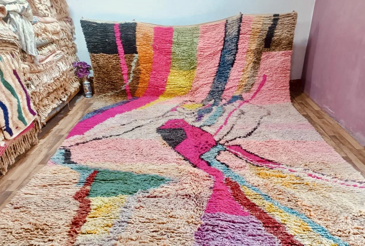 Colorful, abstract-patterned plush rug in a living room with other rugs stacked on a shelf and a small potted plant nearby.