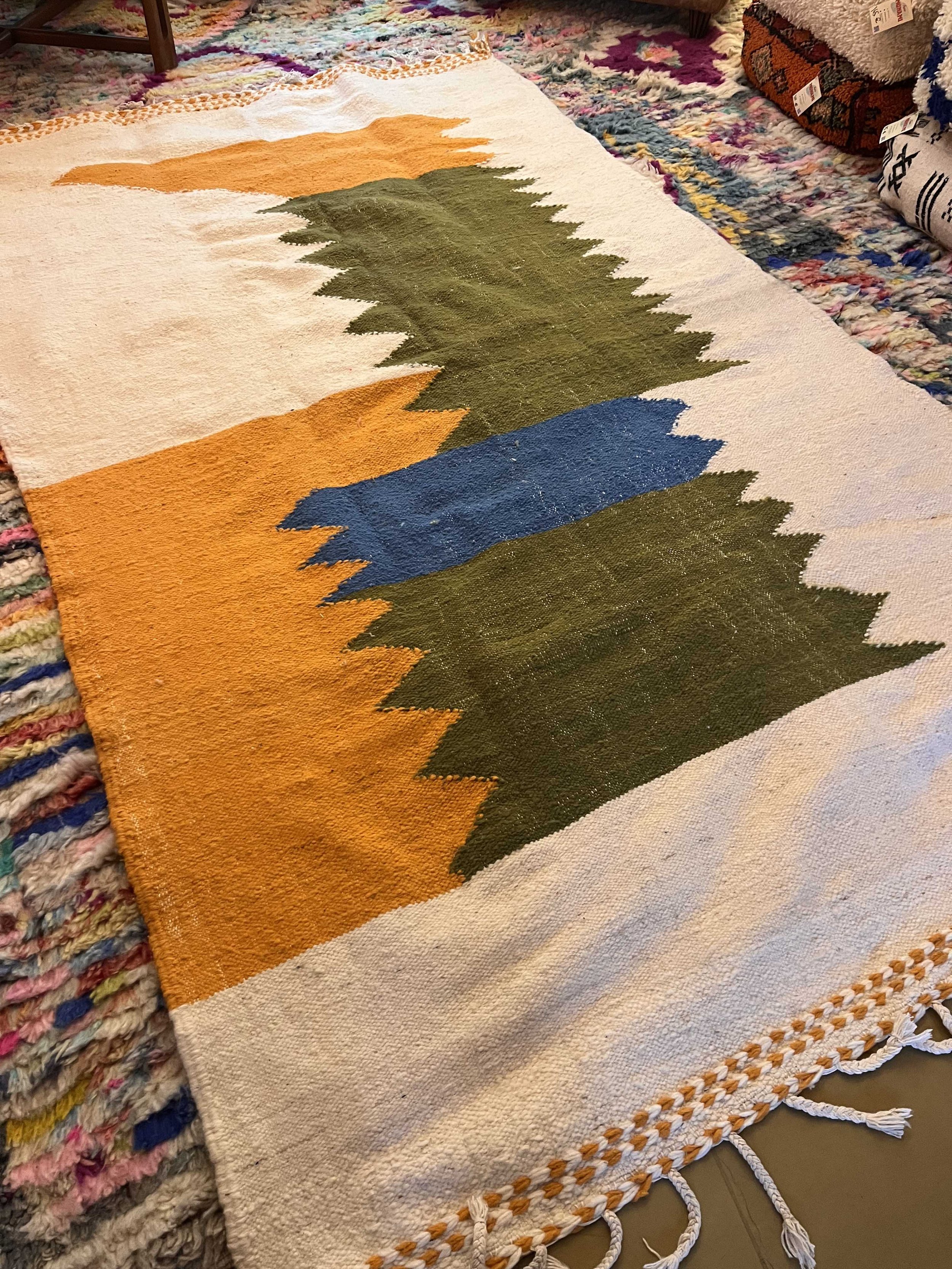 Handwoven woven tapestry with a geometric mountain design in orange, blue, and green, resting on a colorful braided rug.
