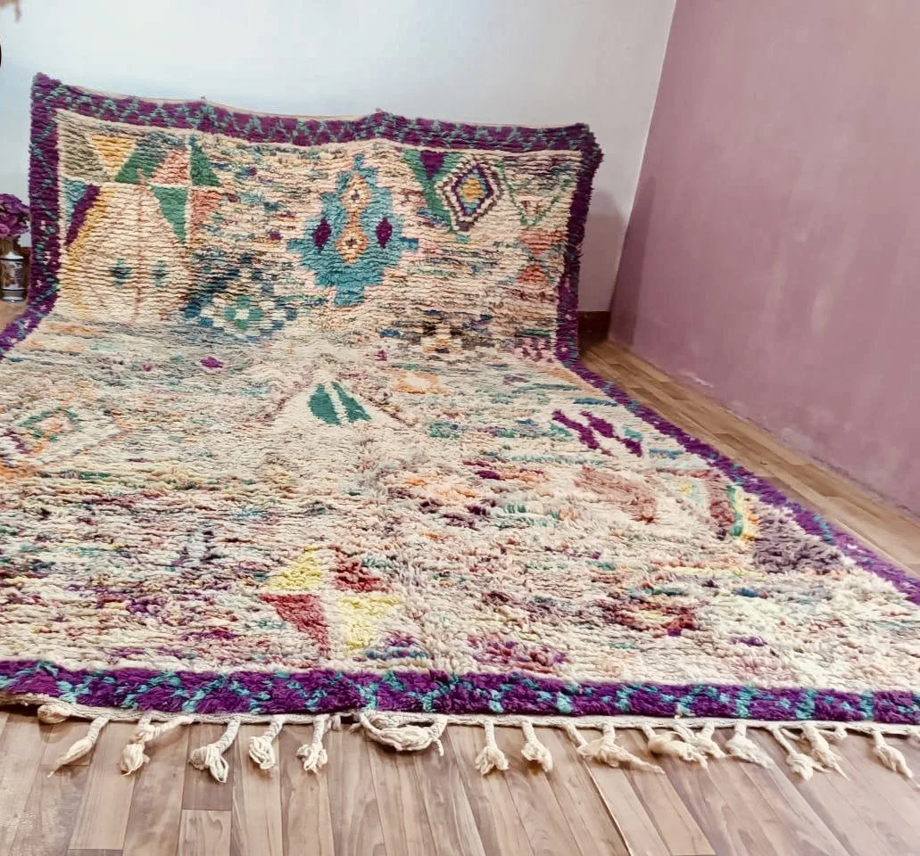 Colorful area rug with geometric patterns and fringe on a wooden floor next to a pink wall and white wall.