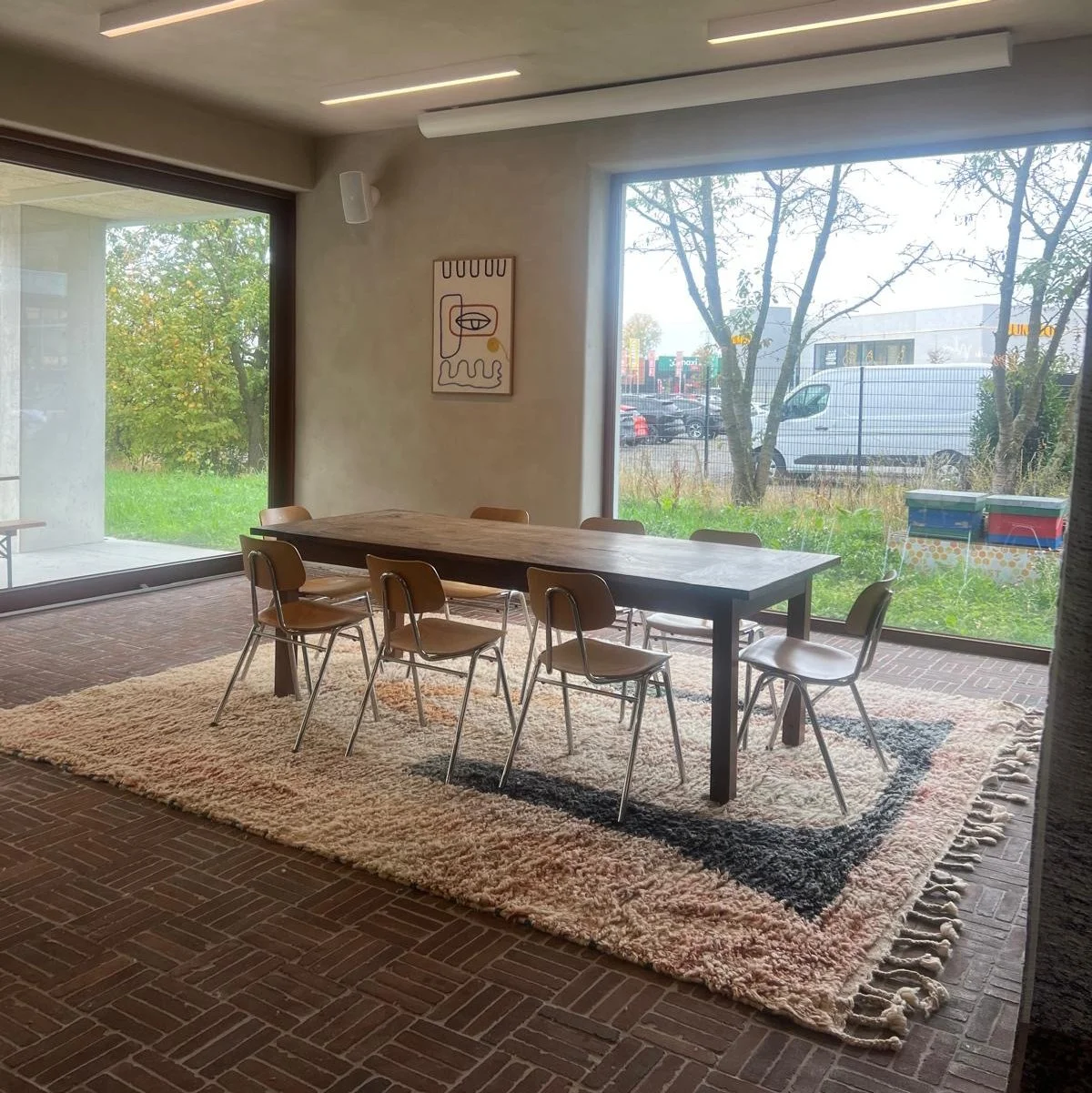 A dining area with a long wooden table and six brown chairs on a large beige and black patterned rug. Large windows reveal trees outside. A piece of abstract wall art is hung on the wall near the window.