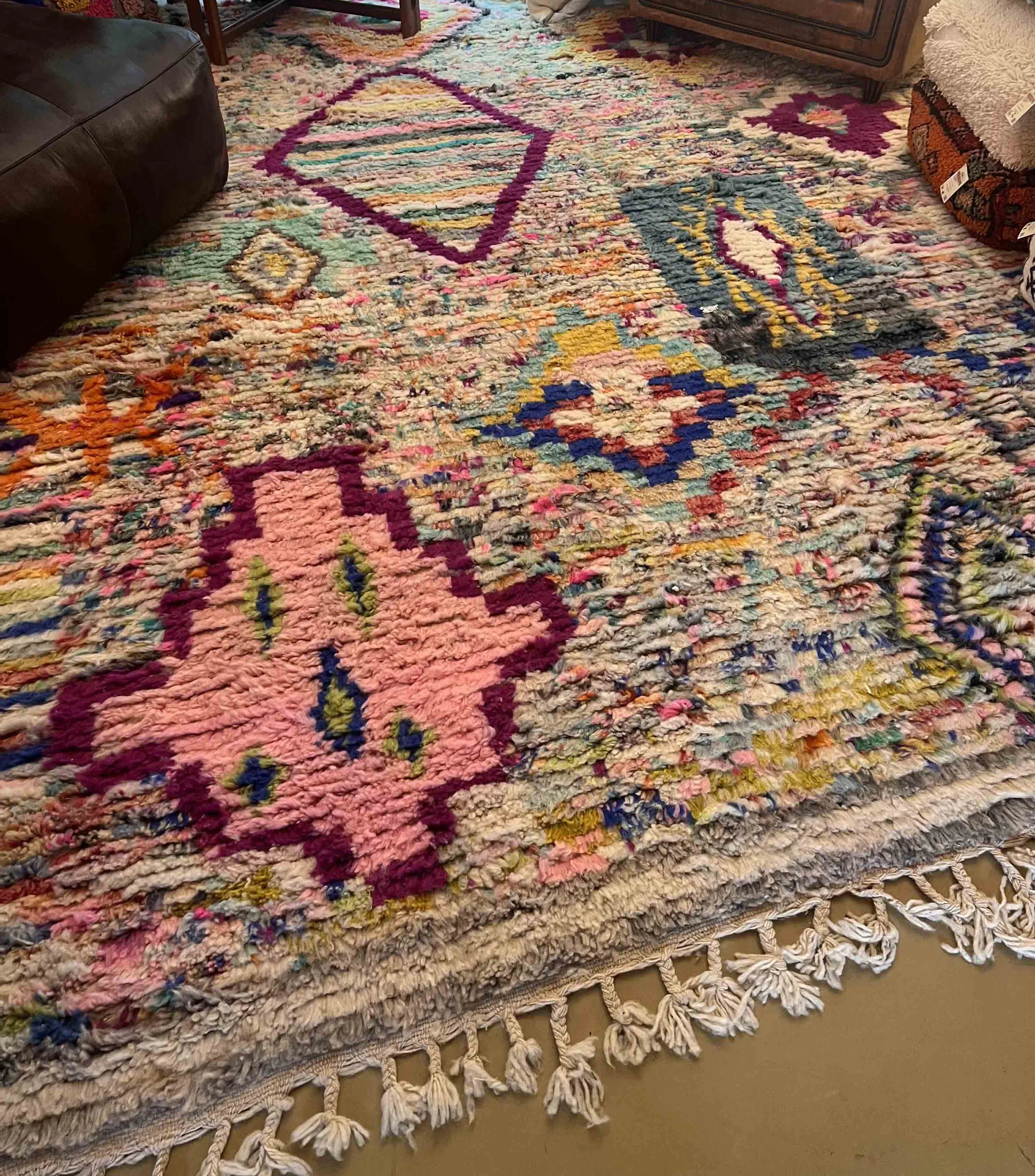 A colorful, patterned rug with various geometric shapes and floral designs, placed on a floor with fringe on the edges. Part of a sofa and other furniture are visible around the rug.