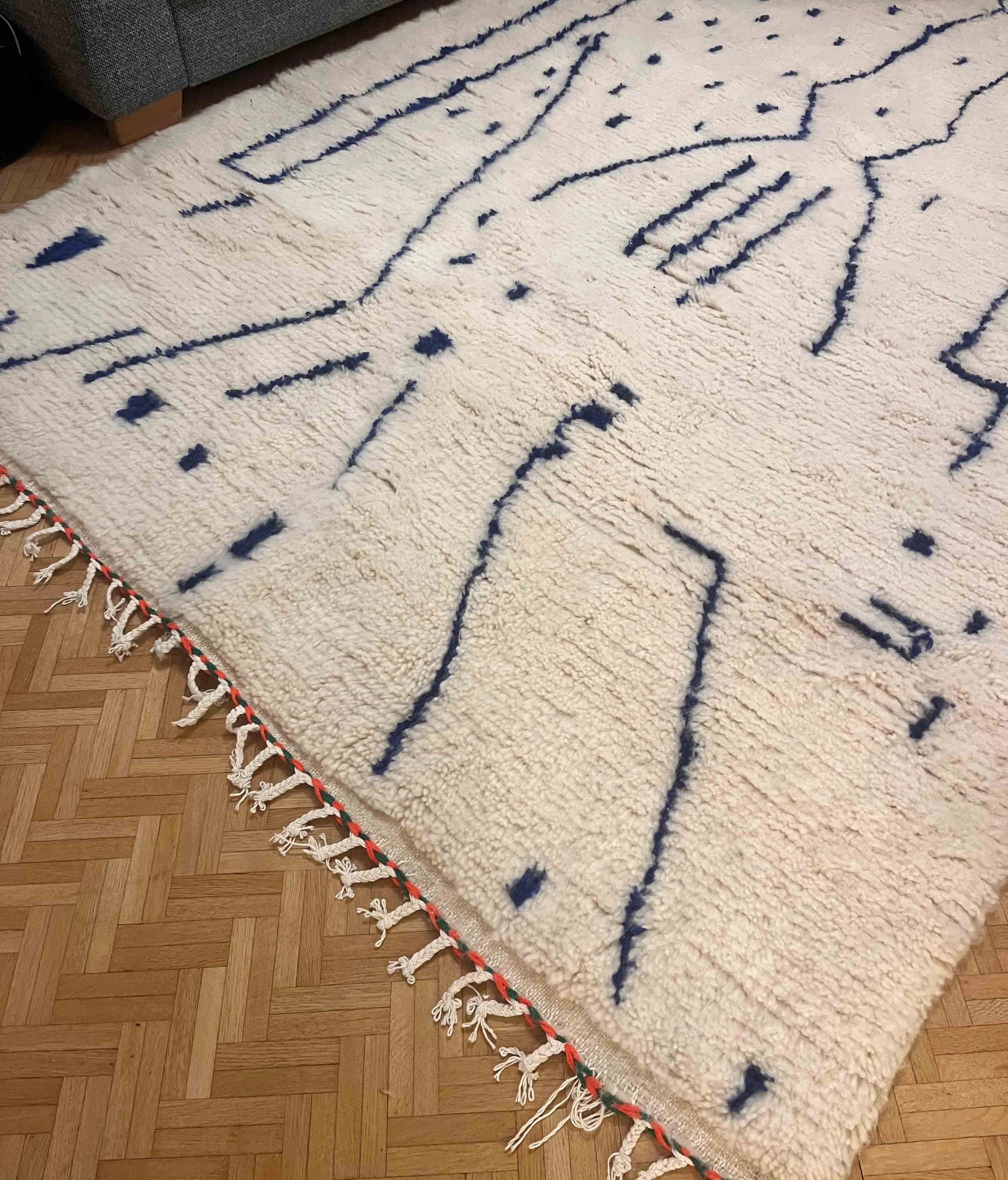 Cream-colored rug with abstract blue lines and dots, fringed border, on a wooden floor near a gray sofa.