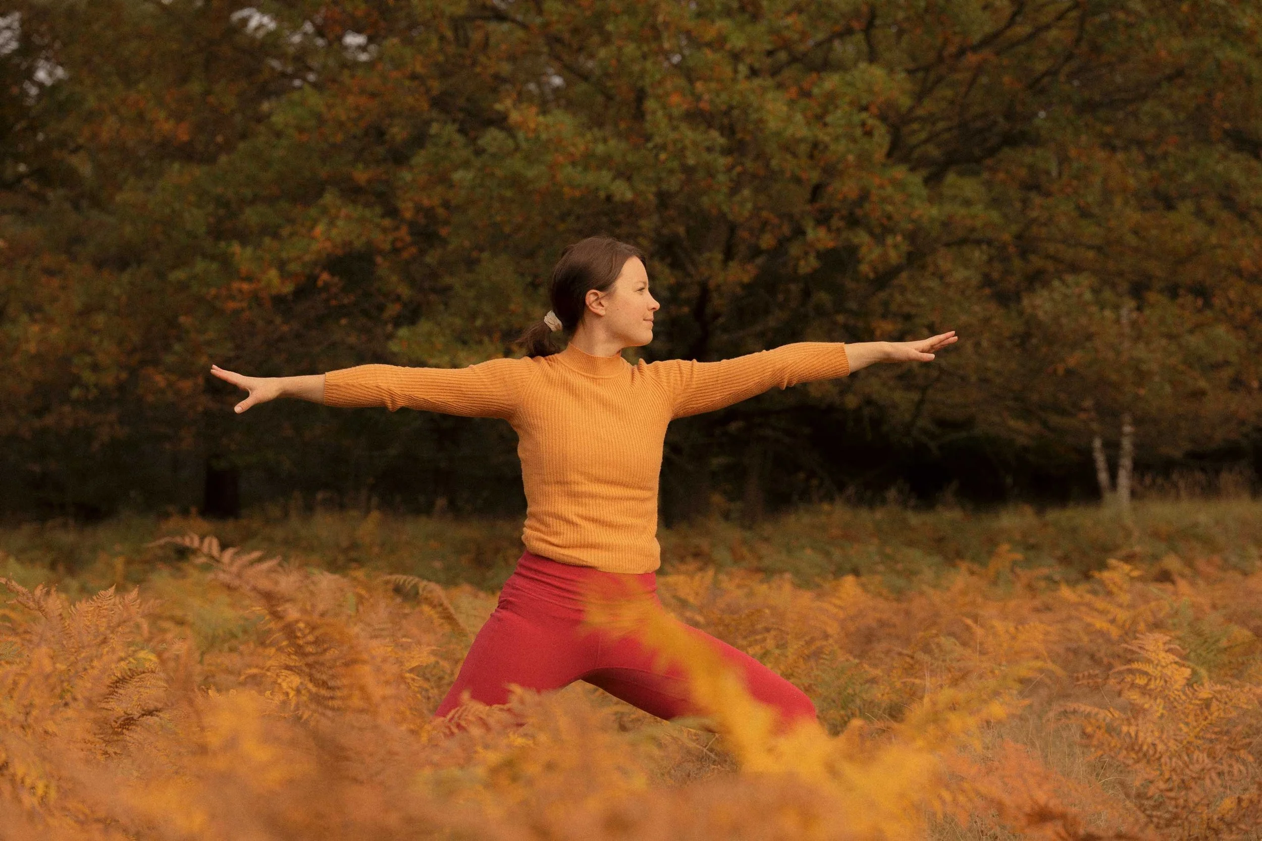 A woman practicing yoga on a dark blue mat on a wooden floor, sitting in a child's pose with her arms extended overhead.