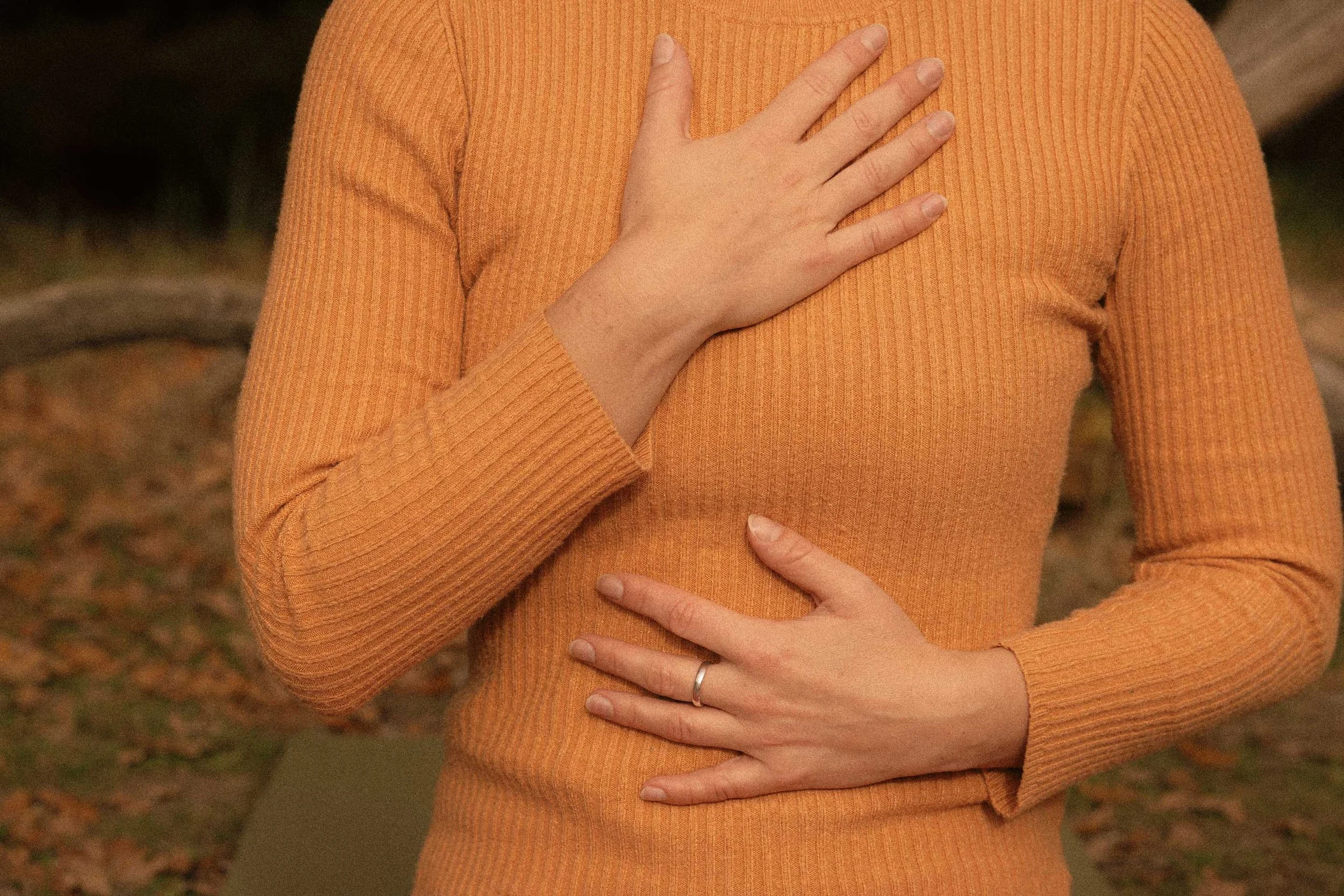 Person wearing an orange ribbed long-sleeve shirt, holding their hand over their chest and abdomen with another hand resting on their stomach.