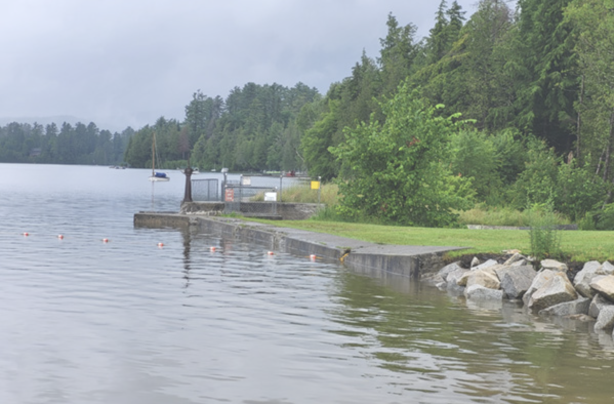 A lakeside scene with a concrete and rocky shoreline, trees, and a boat docked in the water on a cloudy day.