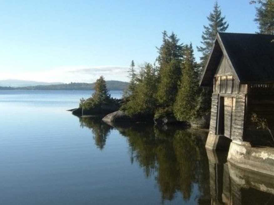 A tranquil lake scene with clear, calm water reflecting the sky and surrounding trees, with a rustic wooden boathouse on the right and a small island with trees in the distance.