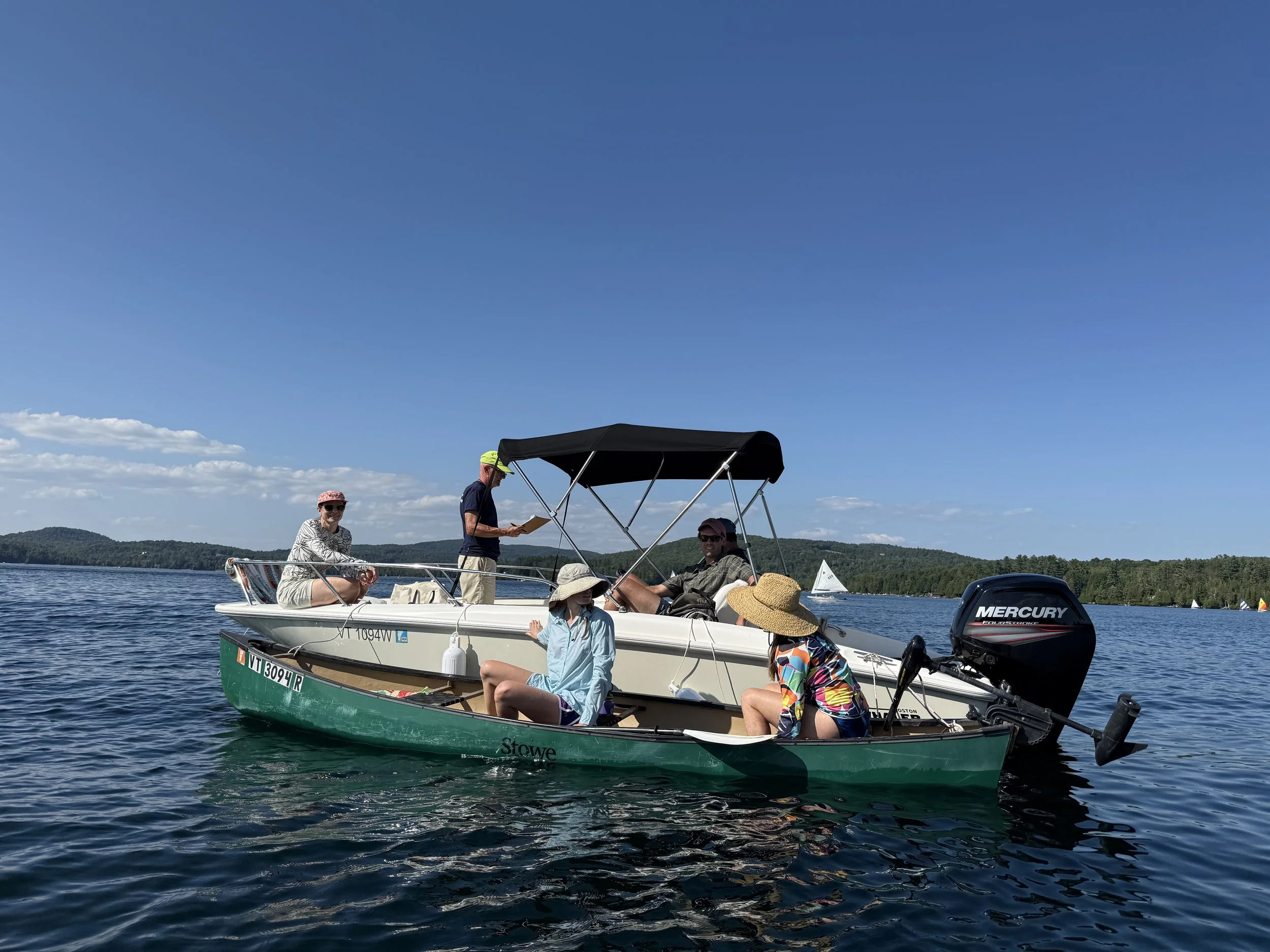 Two boats on a lake with five women and one man enjoying a sunny day. One woman is sitting on the smaller boat, and the others are on the larger boat. The scenery includes calm water, a blue sky, and distant trees.