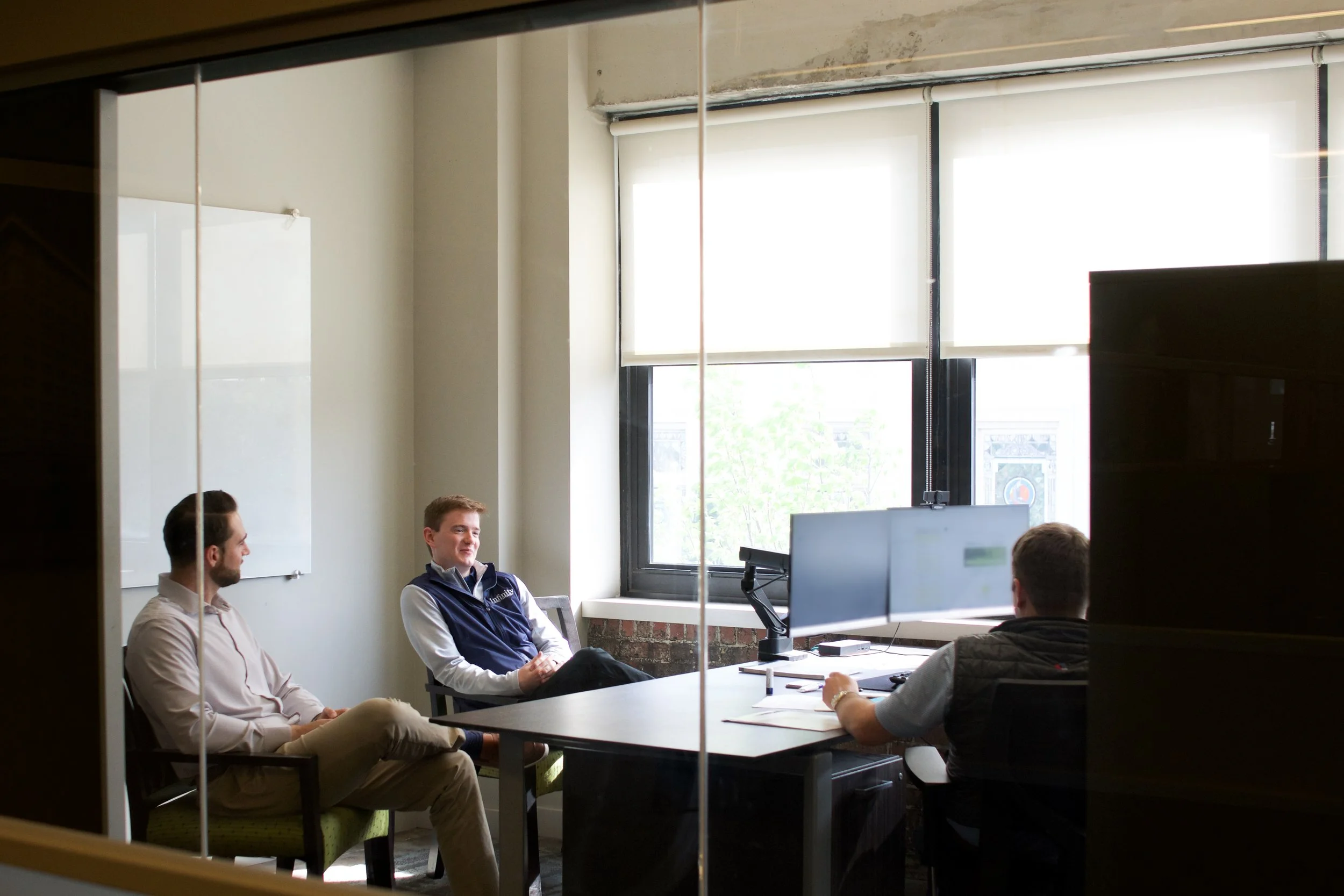 Office employees working at their desks, with one man in a white shirt using a laptop in the foreground and another man in a blue shirt working at his computer in a glass-enclosed office space in the background.