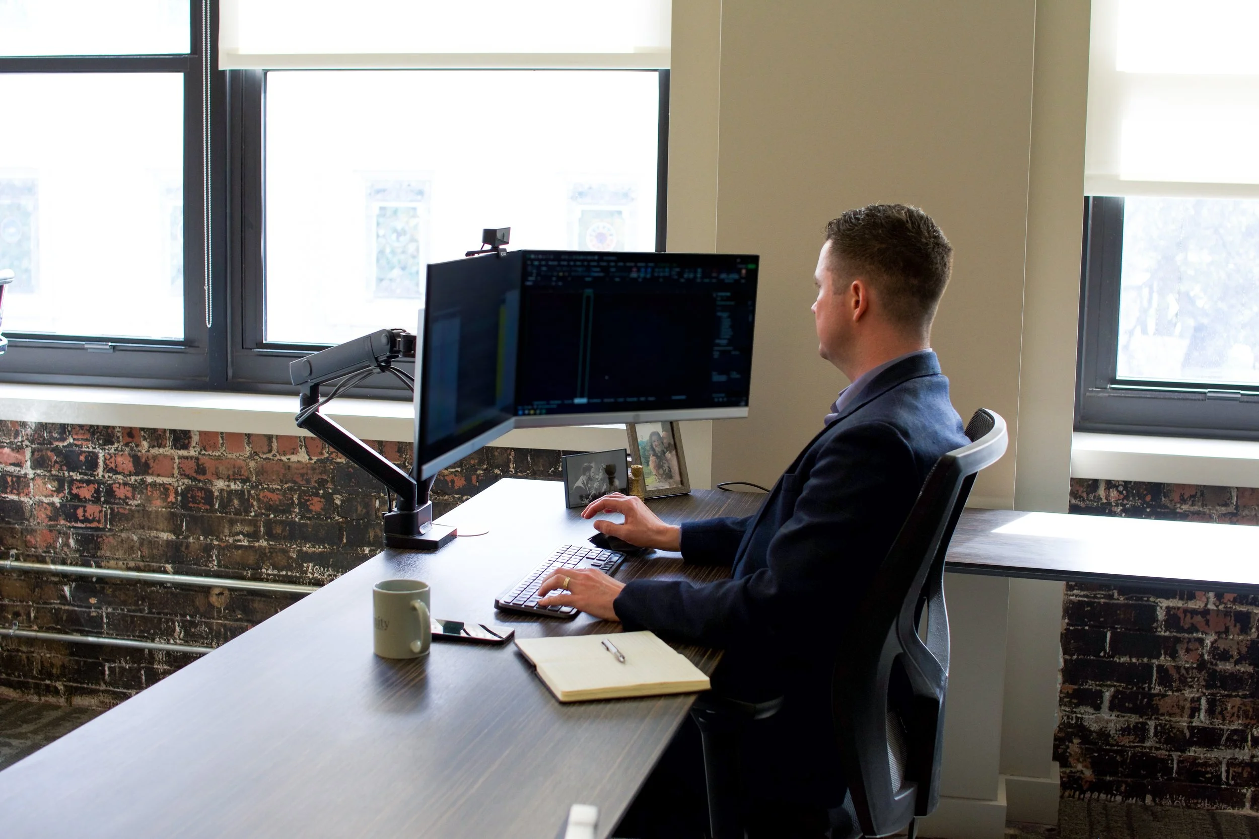 Man working at a desk with dual computer monitors in an office near a window with blinds.