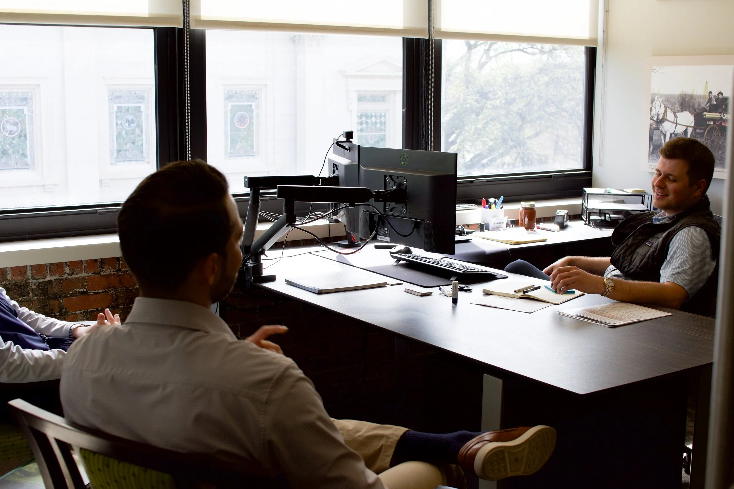 Two men working in an office: one seated at a desk with a computer and the other standing holding a coffee mug.