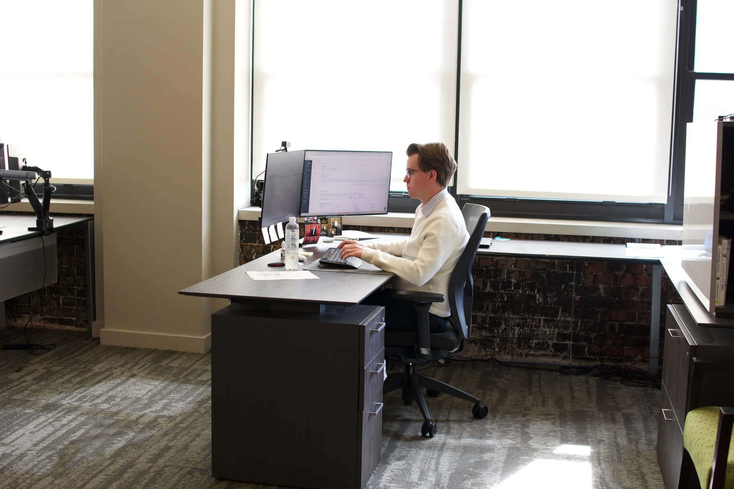 A man sitting at a desk in an office, working on a computer with large windows in the background.