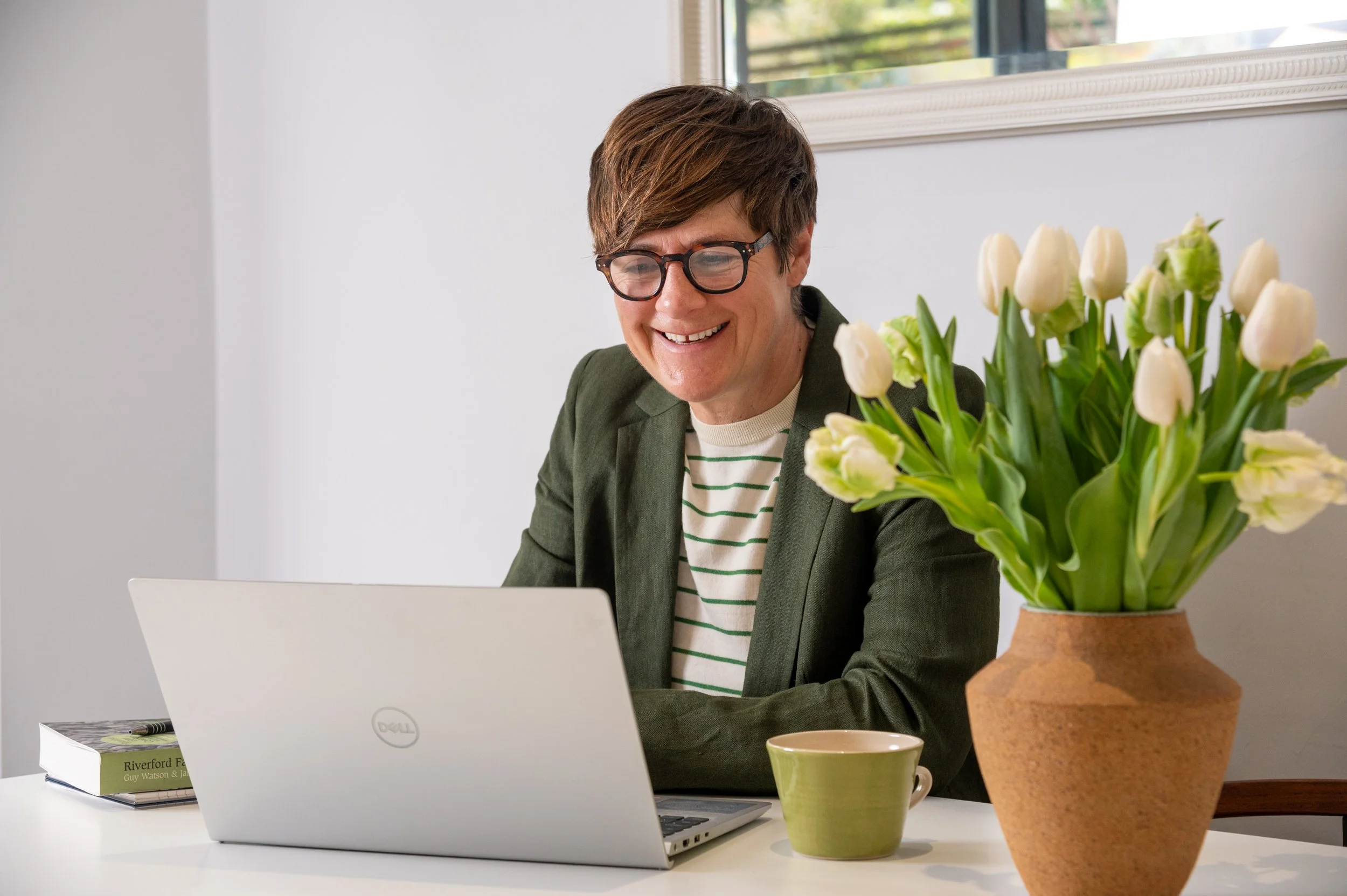 Phillipa with short brown hair, glasses, wearing a green blazer and striped shirt, sitting at a white table with a silver laptop, a green mug, and a book , next to a large terracotta pot of white tulips in a bright room with a window.