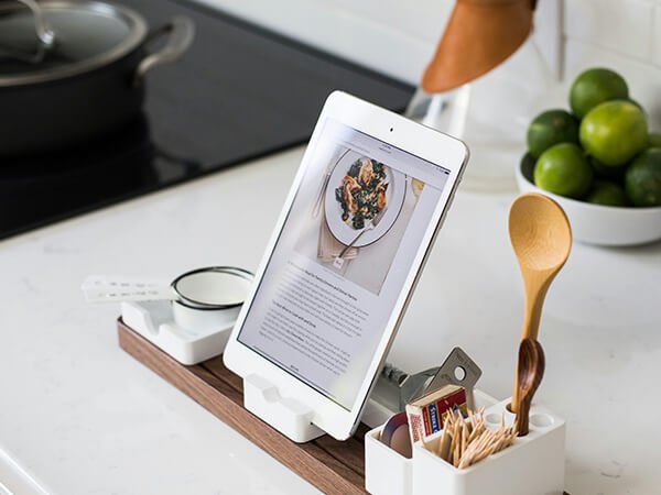 A tablet on a kitchen counter displaying a recipe or food image, surrounded by a spoon, strainer, and condiment packets, with a bowl of limes in the background.