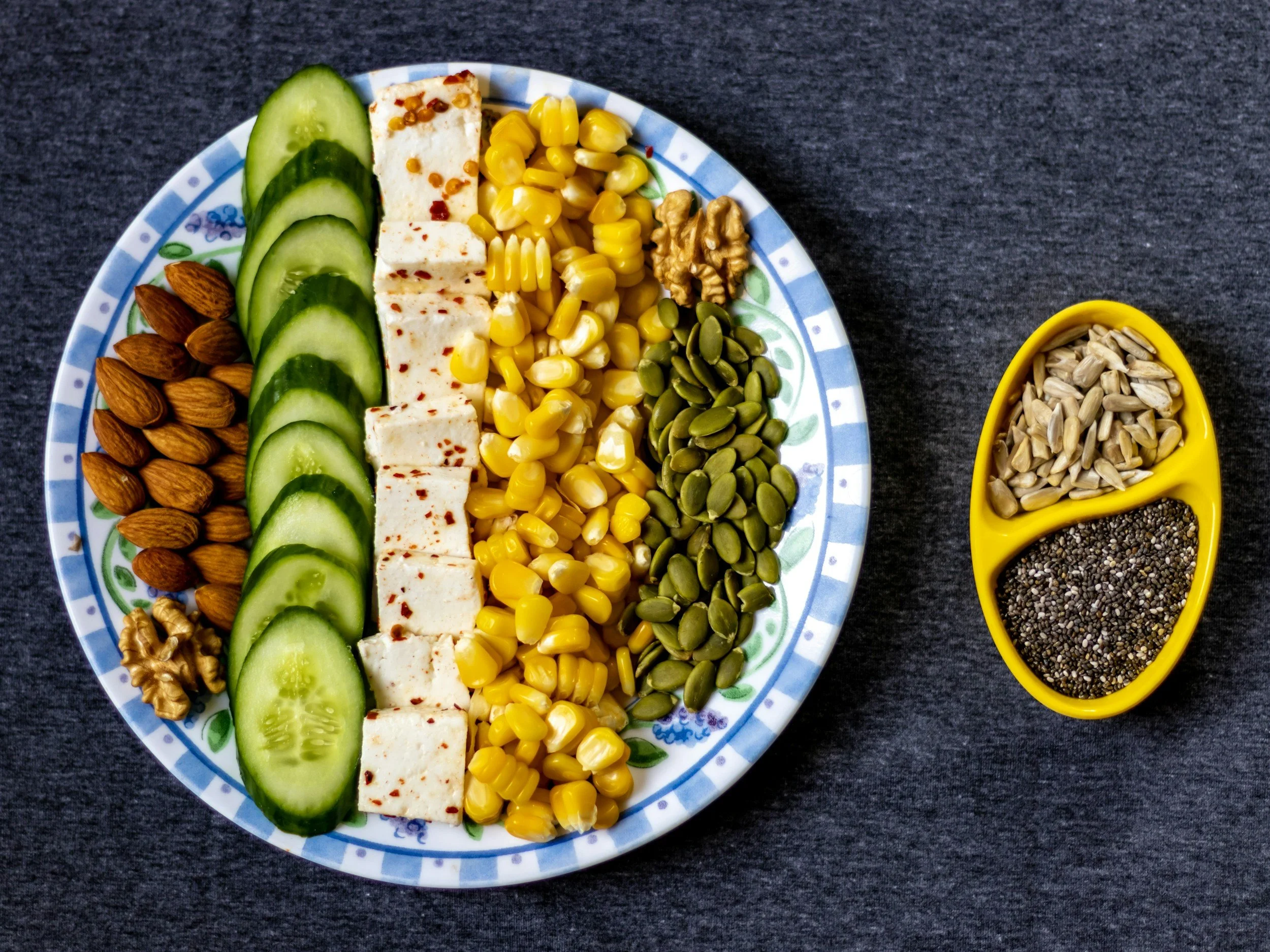 A colorful plate of sliced cucumber, almonds, tofu, corn, walnuts, pumpkin seeds, hemp seeds, and chia seeds on a dark surface with a small yellow dish containing sunflower and chia seeds.