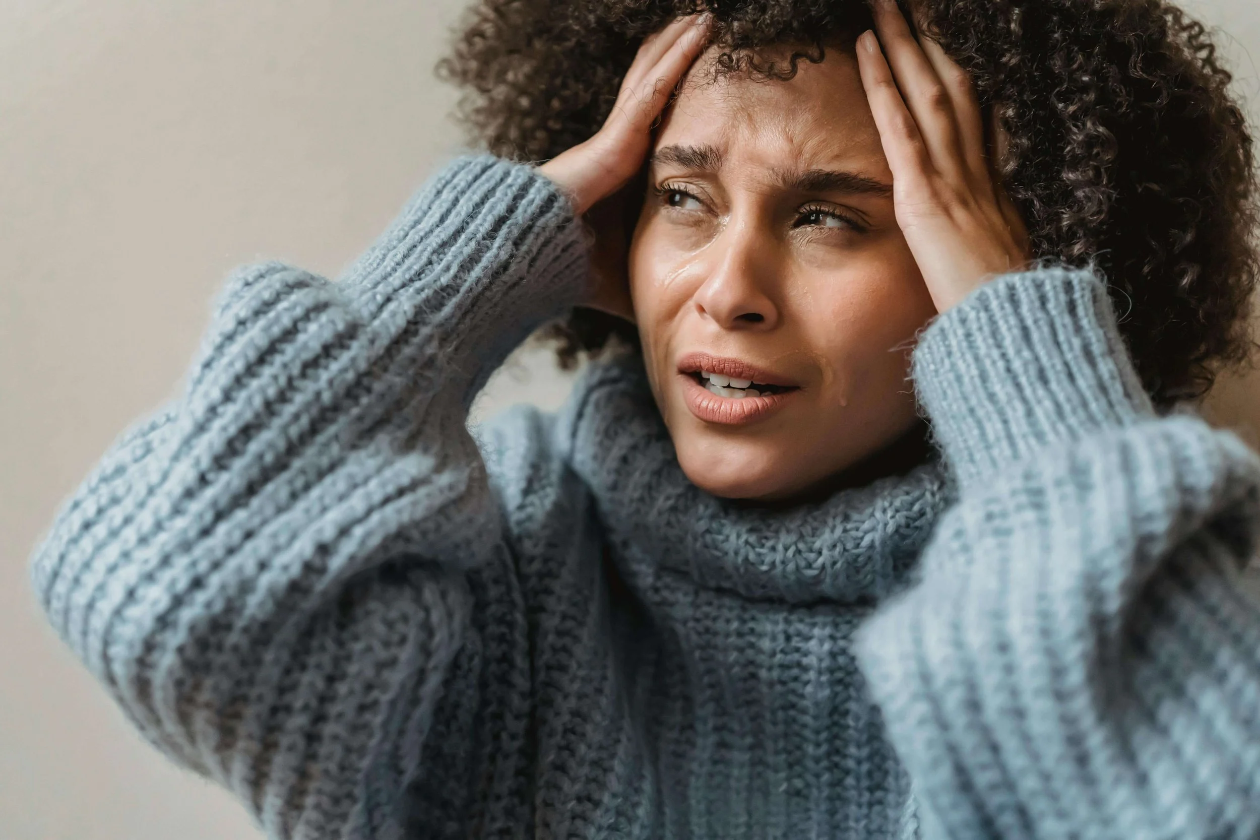 A woman with curly hair wearing a gray sweater, holding her head with both hands, appearing distressed or worried.