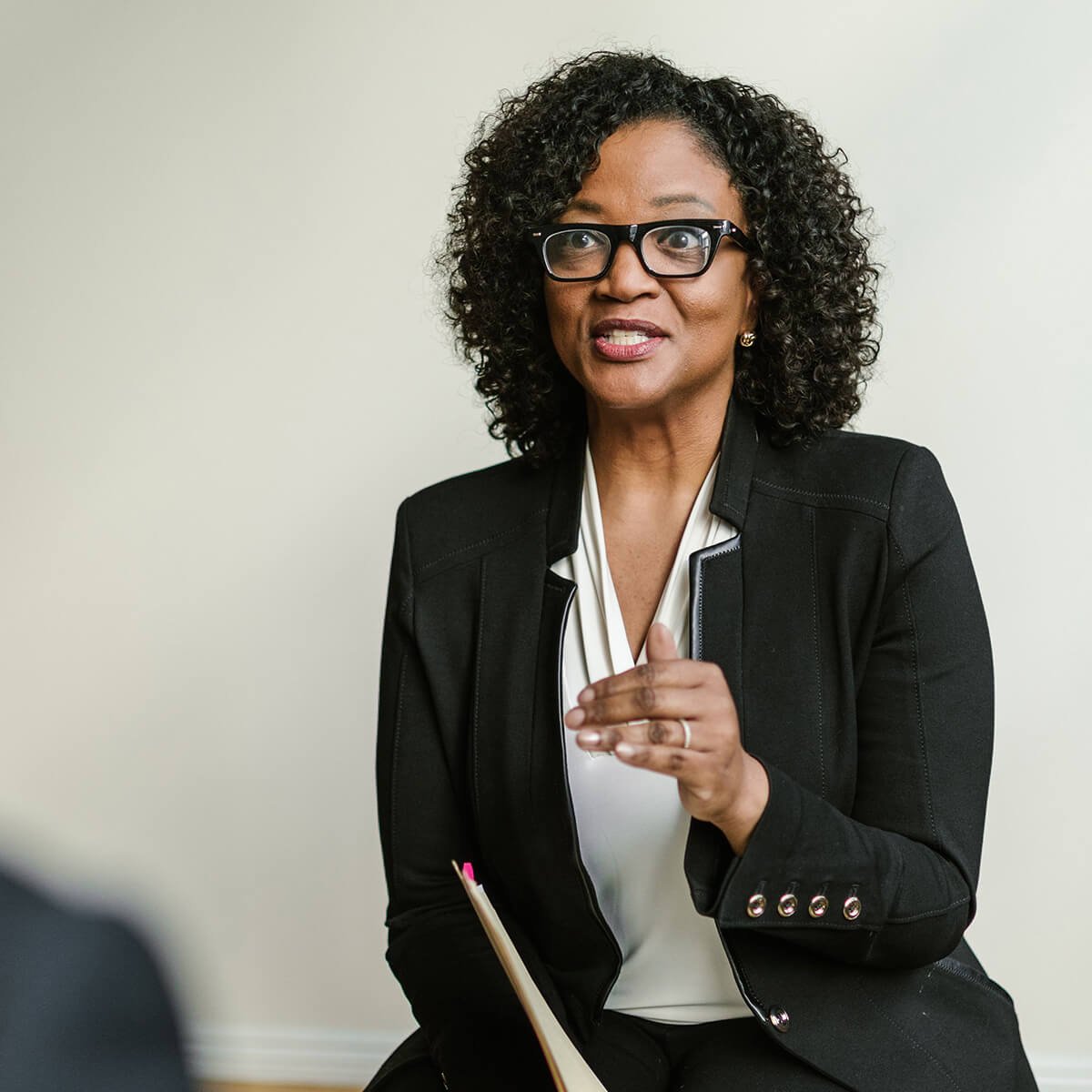 A woman with curly hair and glasses wearing a black blazer and white blouse, sitting and gesturing with her right hand, holding a notebook in her lap, against a plain light-colored wall.