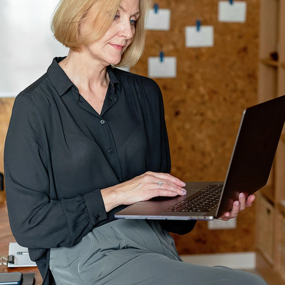 A woman with blonde hair wearing a black blouse sitting and looking at her laptop in an office setting.