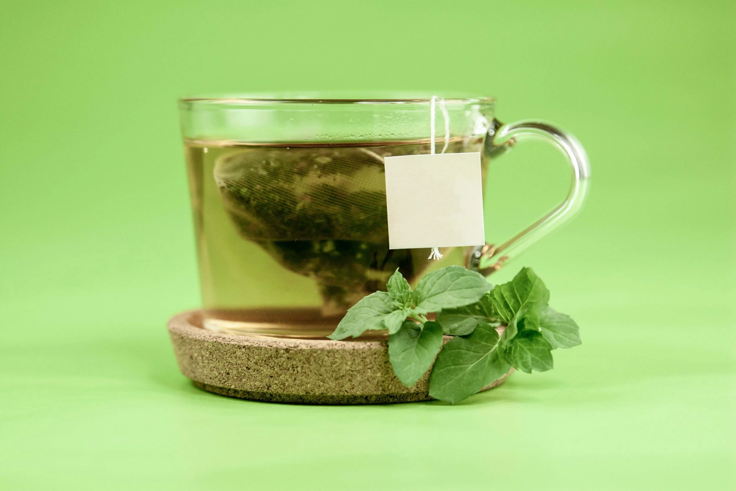 A glass teacup with tea and a tea bag inside, placed on a small round cork coaster. Fresh green mint leaves are positioned underneath the coaster. The background is a solid bright green color.