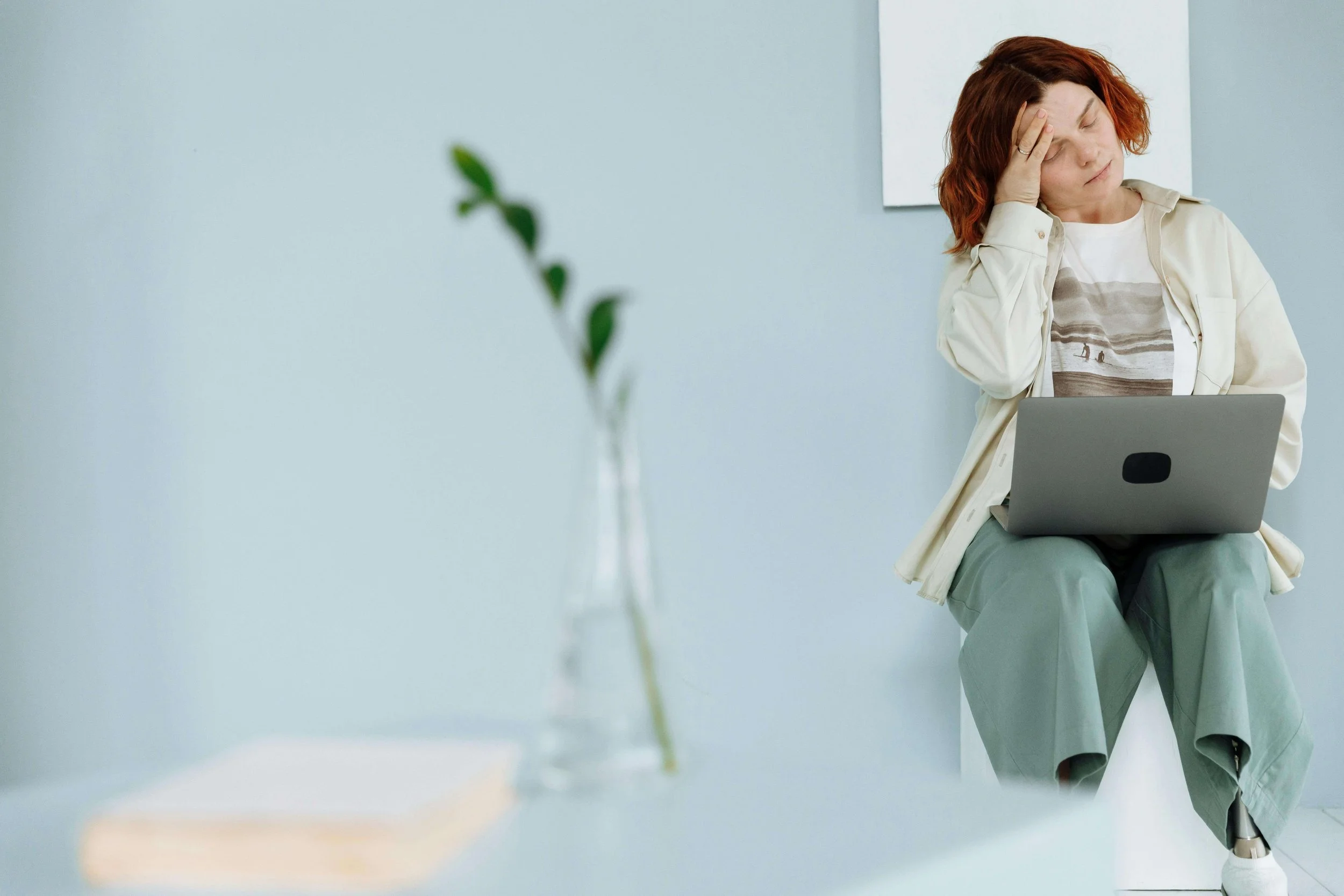 Woman sitting with laptop, holding head in a minimalist space, depicting anxiety and hot flush.