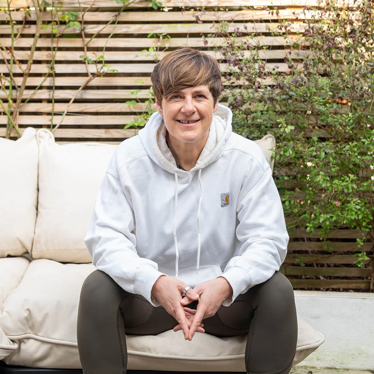 Phillipa Jacobs-Smith smiling with short brown hair sitting outside on a cream cushion sofa, wearing a white hoodie and green leggings, with a wooden slatted fence and green plants in the background.
