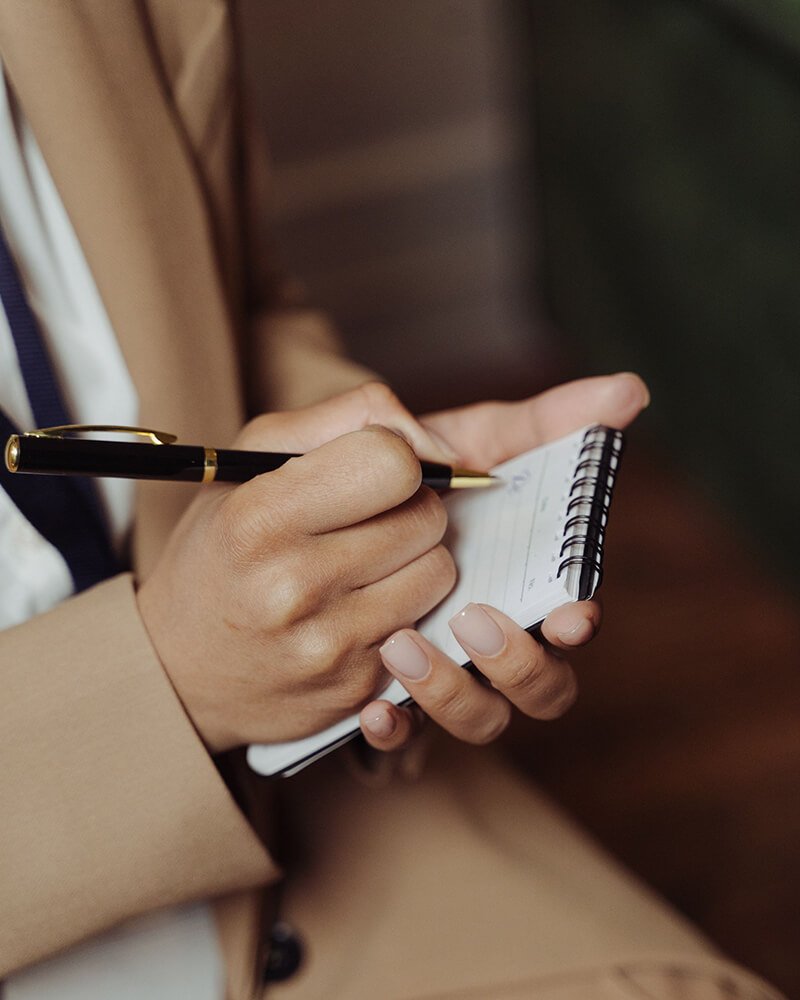 A person with light skin writing in a small spiral notebook with a black and gold pen, wearing a beige coat.