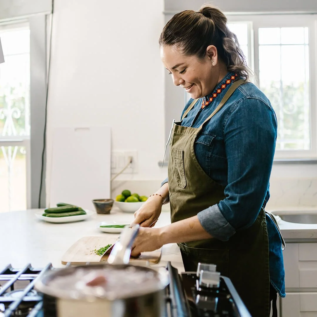 Woman with dark hair in a ponytail, smiling and chopping vegetables in a kitchen, wearing a denim shirt and an apron.