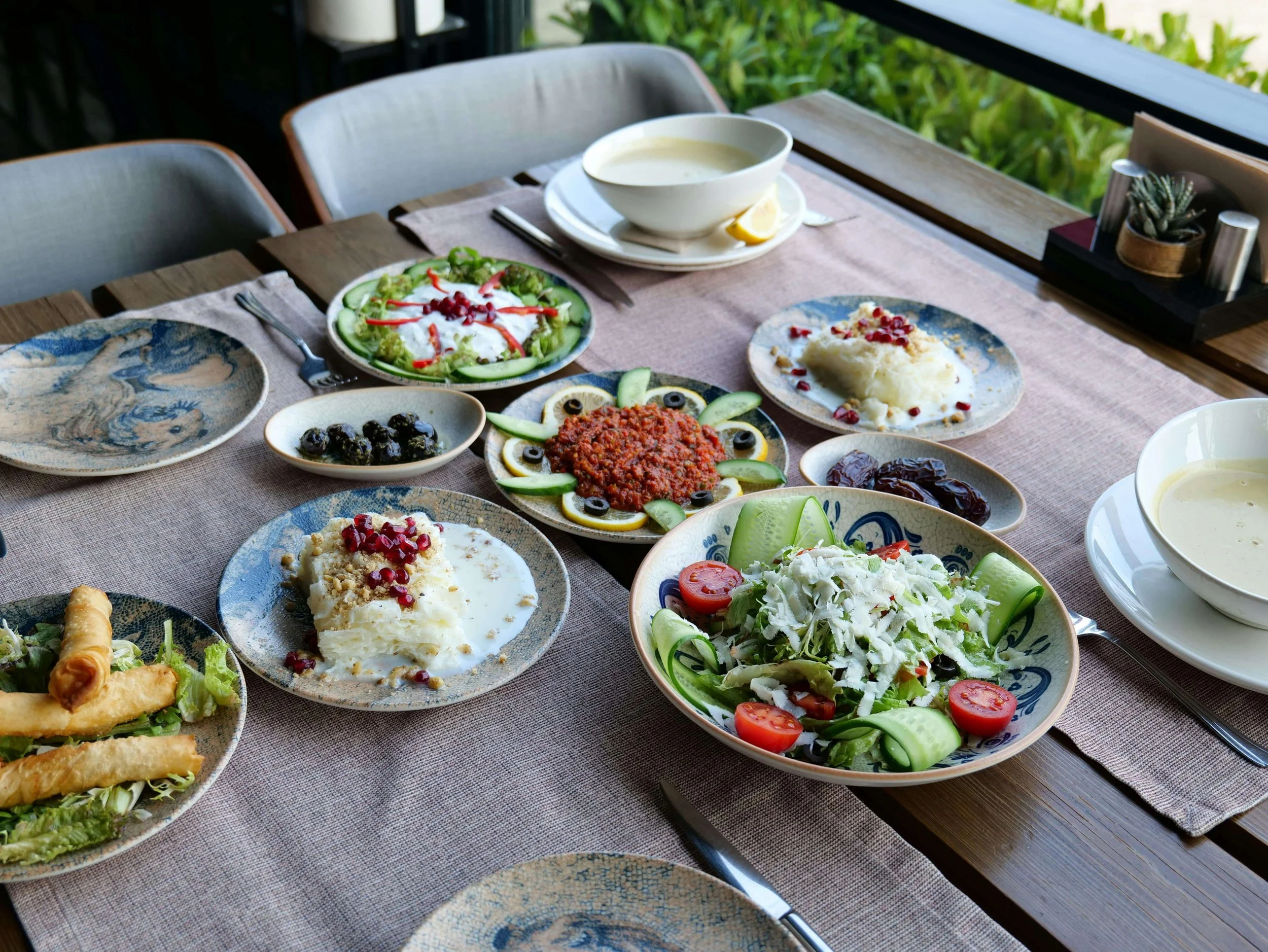 A table set with various dishes on ceramic plates and bowls, including salads, mashed potatoes with pomegranate seeds, a bowl of soup, and a plate of pita bread, on a beige tablecloth outdoors near green foliage.