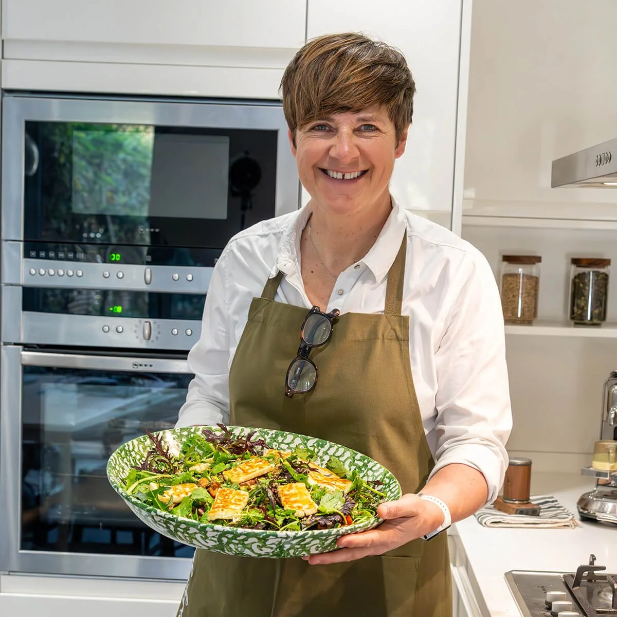 Smiling woman in a white shirt and brown apron holding a plate of salad in a modern kitchen.