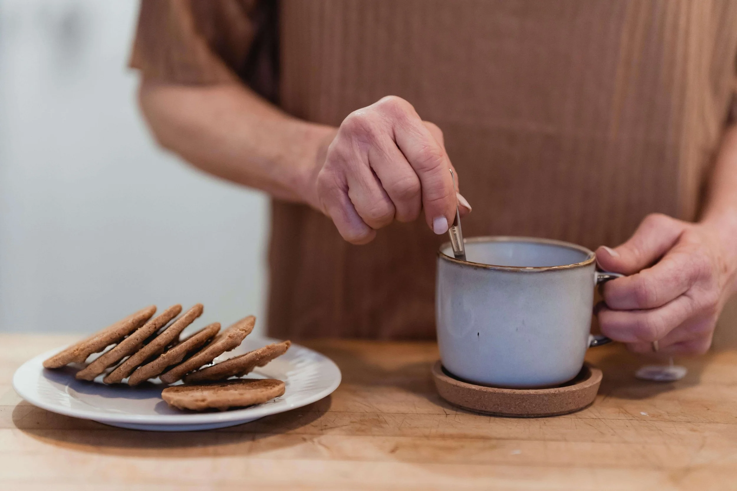 A person’s hand holding a spoon over a white mug on a cork coaster, with cookies on a plate in the foreground.