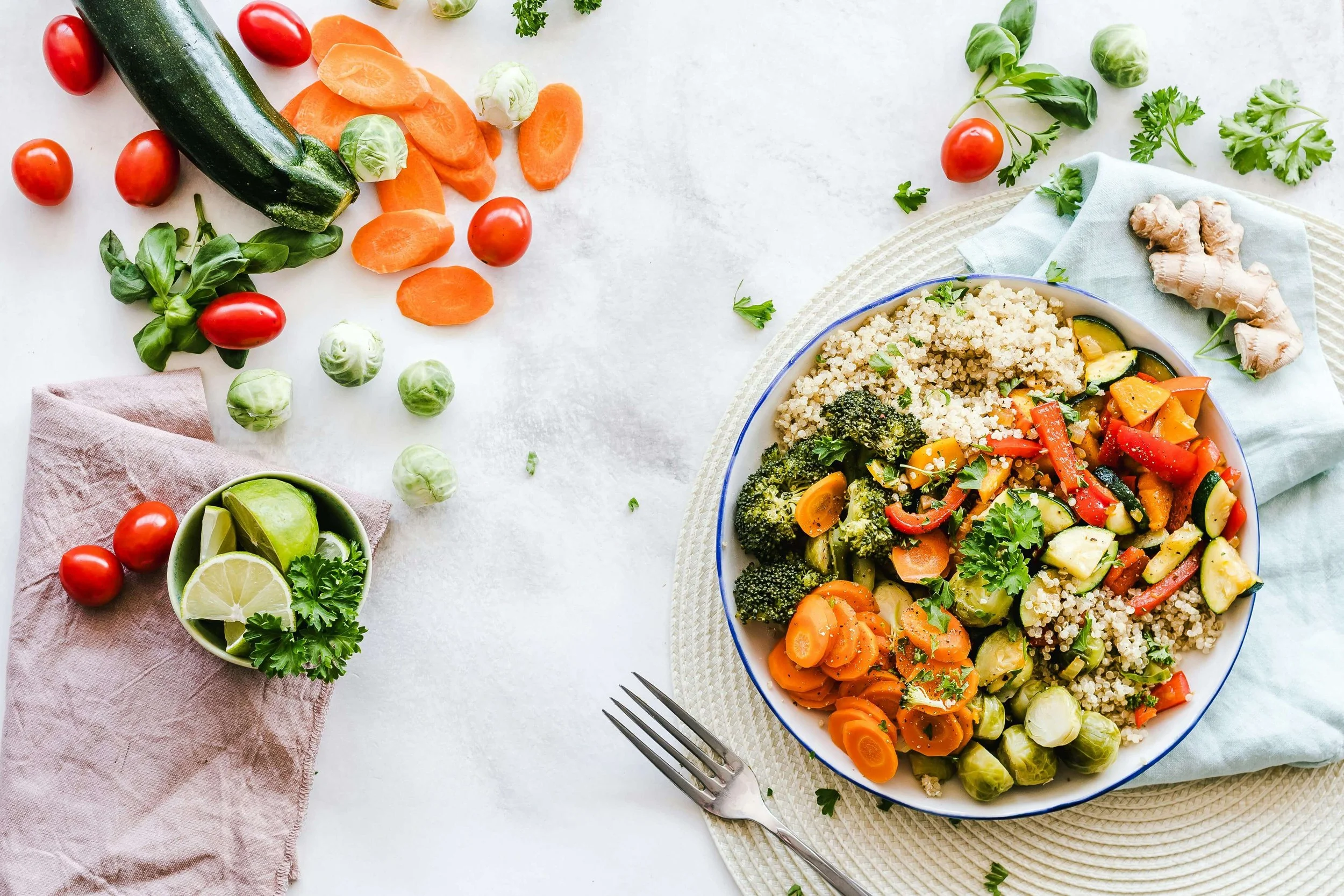 Fresh vegetables including zucchini, cherry tomatoes, brussels sprouts, carrots, ginger, lemon, lime, and herbs arranged on a white surface near a bowl of cooked vegetable salad with quinoa.
