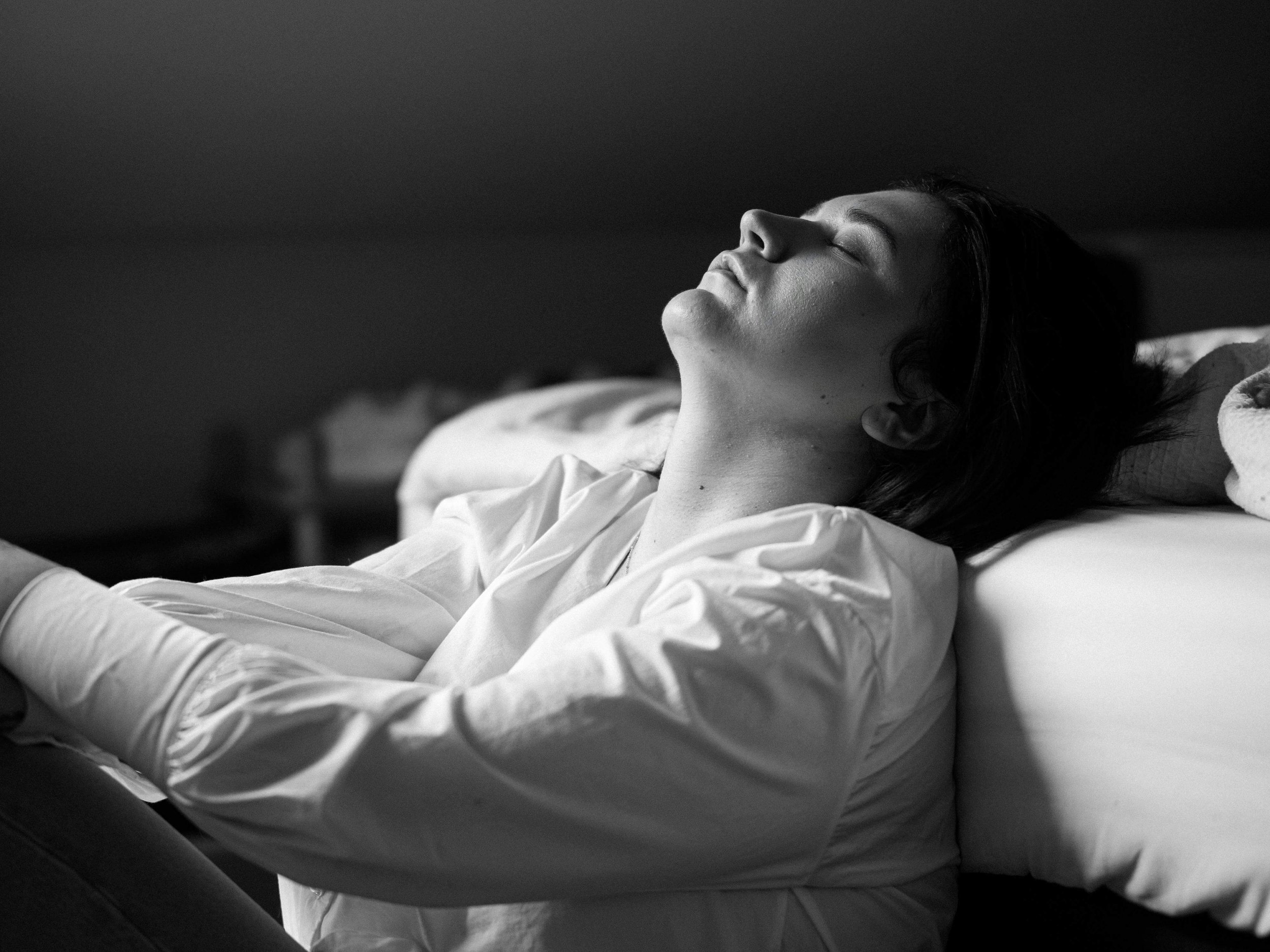 Fatigued woman in black and white, eyes closed, sitting by a bed in a tranquil indoor setting.