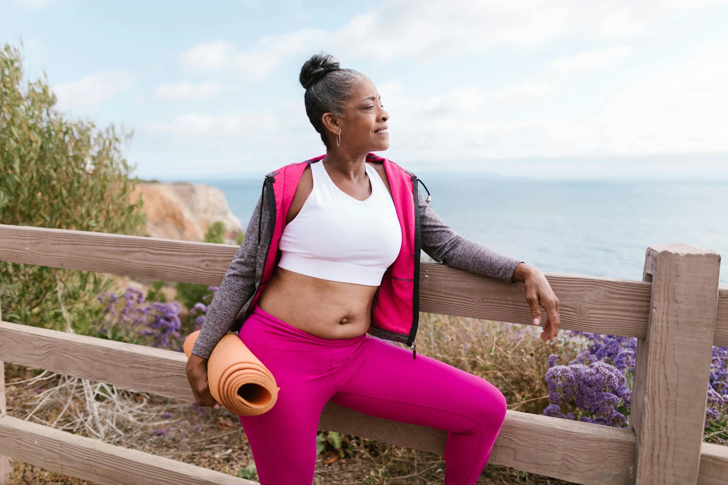 Older black woman in yoga clothing leaning against a fence overlooking the beach.