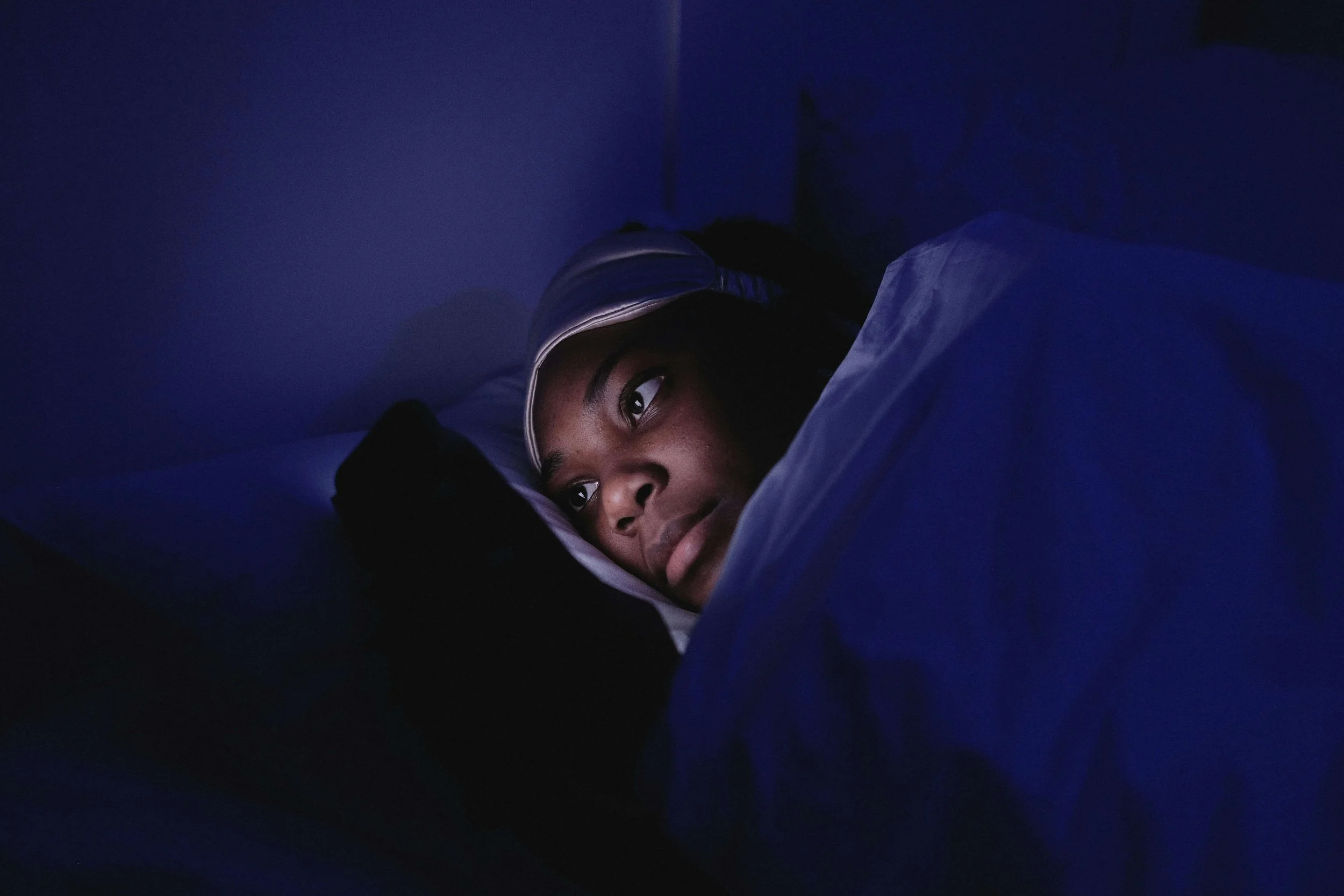 A person lying in bed with dark lighting, wearing an eye mask on her forehead, awake when trying to be asleep.
