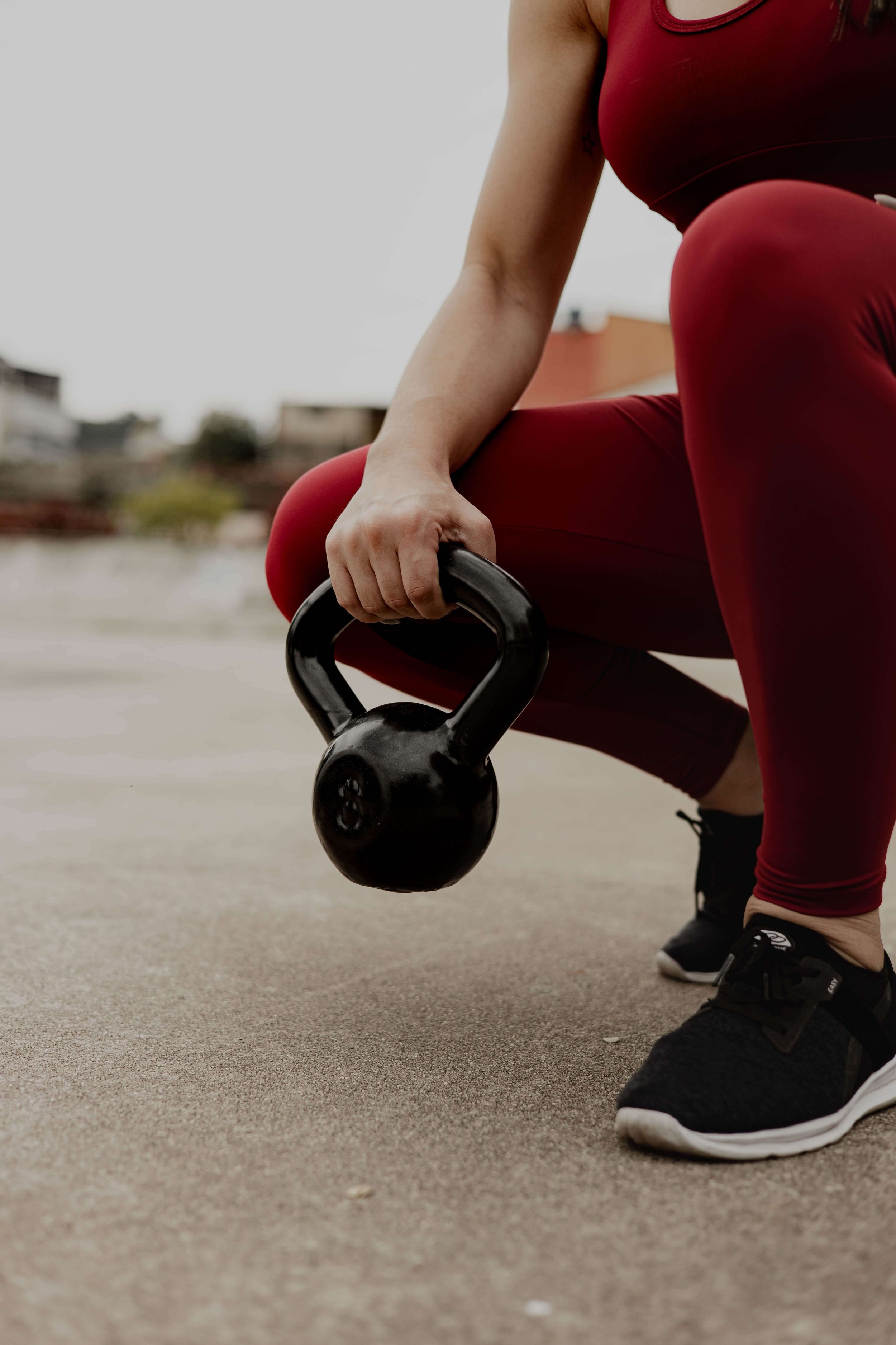 A woman in red workout leggings and black trainers outside crouching with a kettle bell in her hand