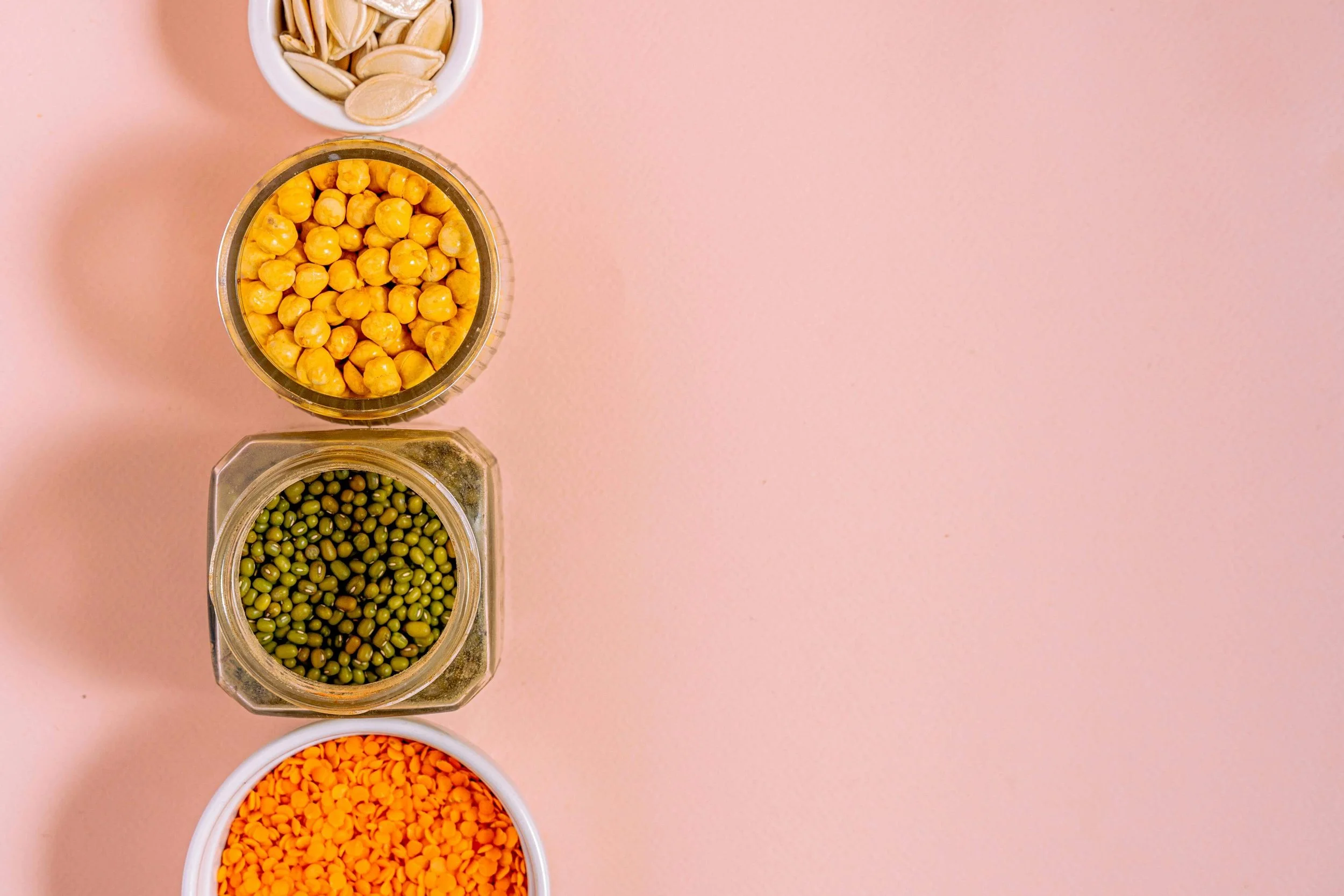 Top view of four small bowls containing different colored dry foods on a pink background.