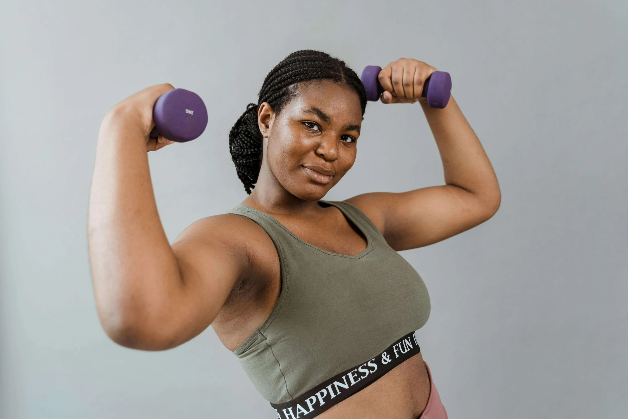 A young woman with dark braided hair wears a green sports bra and pink leggings, lifting purple dumbbells and smiling.