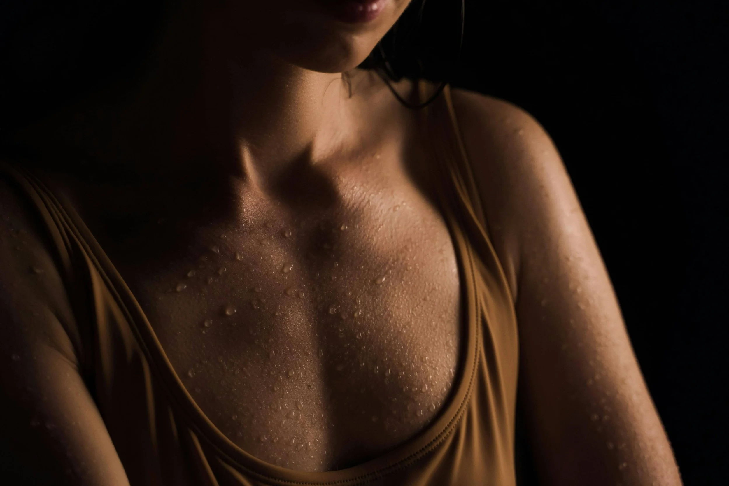 Close-up of a woman's face and shoulder, with water droplets on skin, wearing a sleeveless top, in low lighting.
