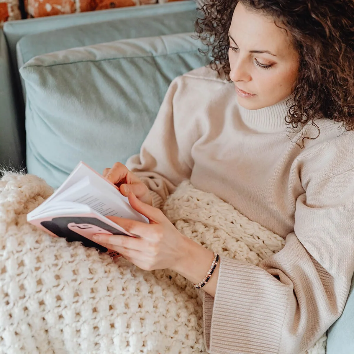 Woman with curly hair wearing a beige sweater, sitting on a couch, reading a book, with a cream-colored knit blanket on her lap.