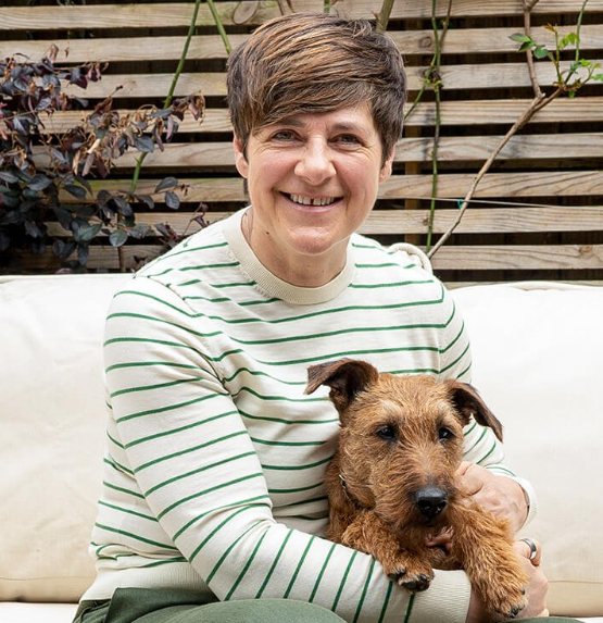 A woman with short brown hair smiling while sitting on a white couch, holding a small brown dog with a scruffy coat, in front of a wooden fence with plants.