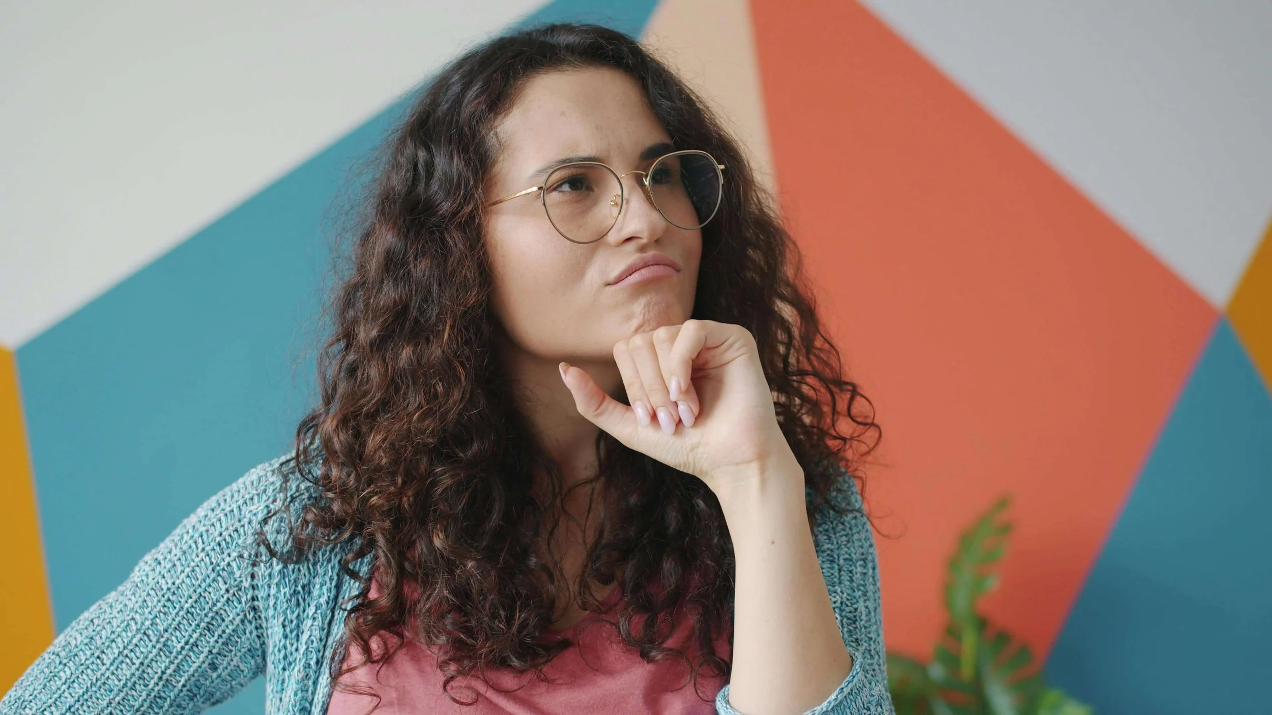 A woman with curly brown hair and glasses resting her chin on her hand in front of a colorful geometric wall.