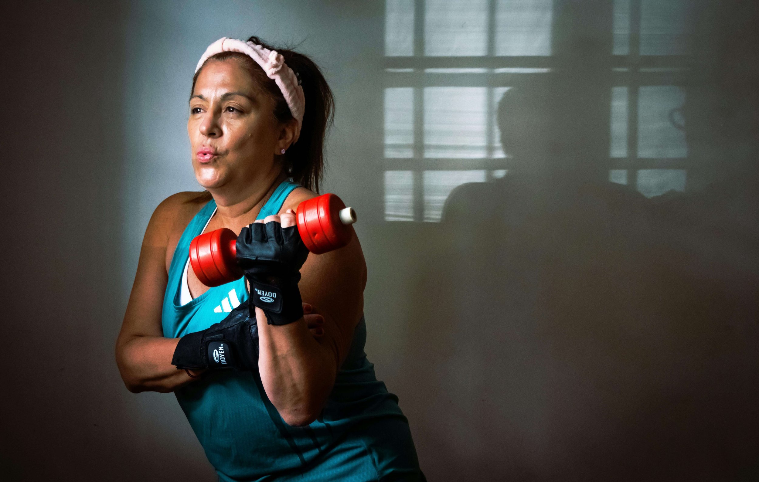A woman with red hair wearing a pink headband and athletic clothing, holding a red dumbbell, standing in front of a window with a shadowed reflection.