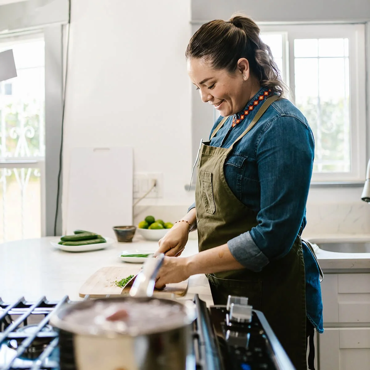 A woman in a denim shirt and green apron is chopping vegetables in a bright kitchen with white cabinets and large windows.
