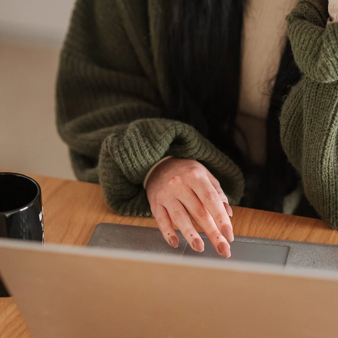A person with long dark hair wearing a greenish-brown oversized knit sweater working on a laptop on a wooden desk, with a black mug nearby.