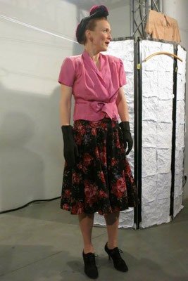 Frau in Vintage-Kleidung mit pinkem Top, schwarzer Glitzerskirt, schwarzen Handschuhen und Hut, stehend in einer Galerie.