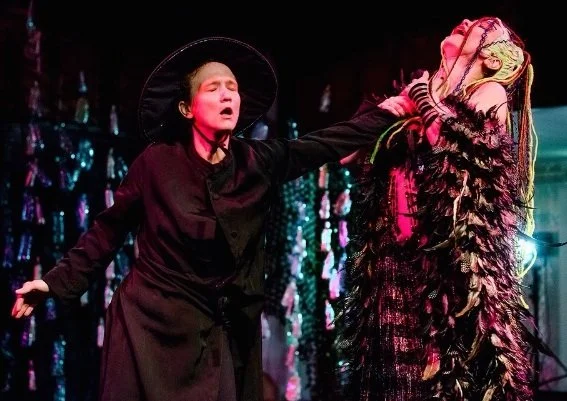 Zwei Frauen in auffälligen Kostümen auf einer Bühne bei einer Aufführung, eine greift nach der anderen.