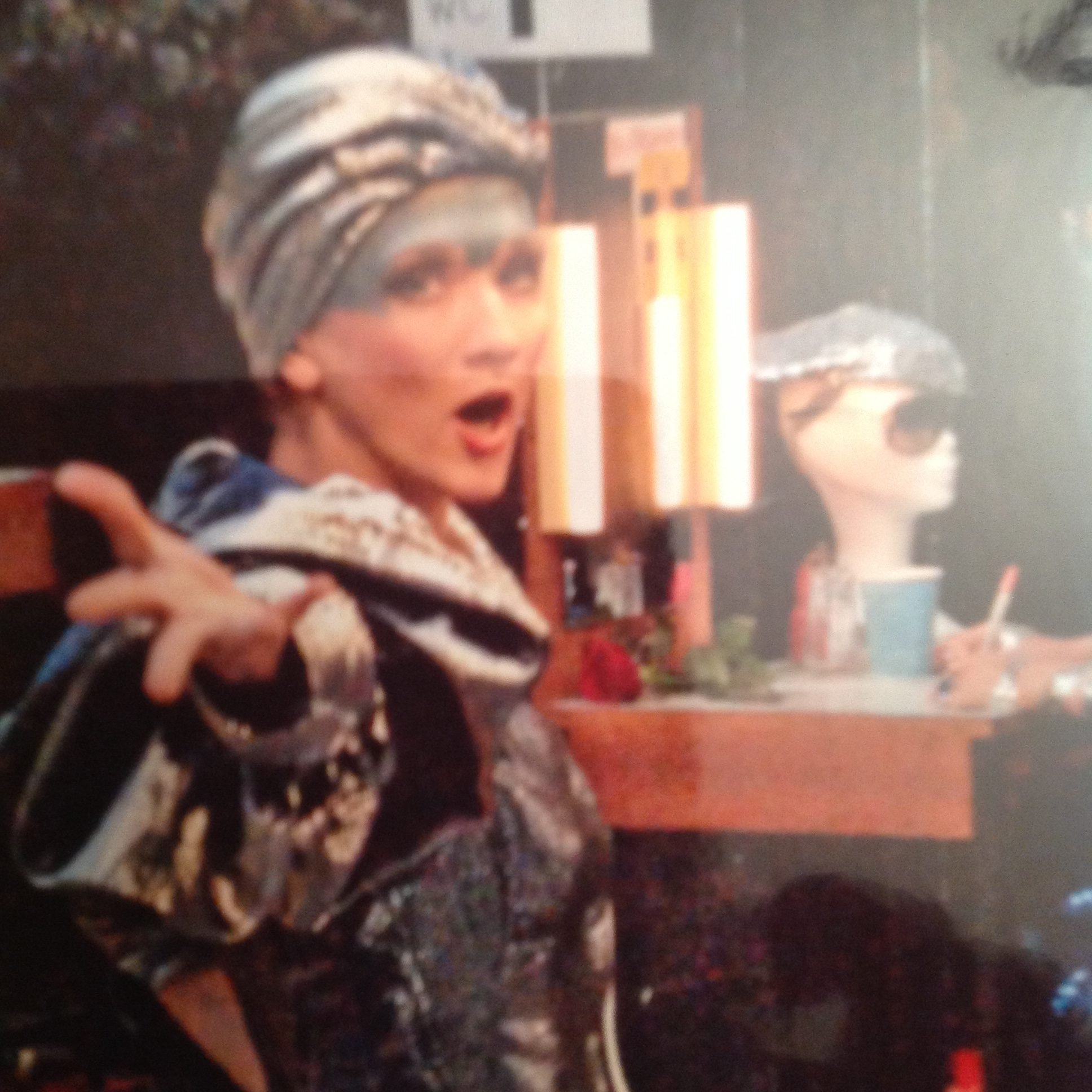 Frau mit einer gestreiften Kopfbedeckung und auffälligem Make-up, die überrascht schaut, im Vordergrund. Im Hintergrund eine Statue und eine Tisch mit einer blauen Tasse und einem Stift.