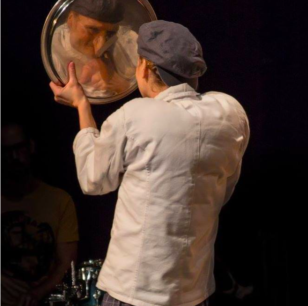 Ein Mann in einer weißen Kochjacke und einer schwarzen Kochmütze schaut in einen runden Spiegel und sieht sein eigenes Spiegelbild.