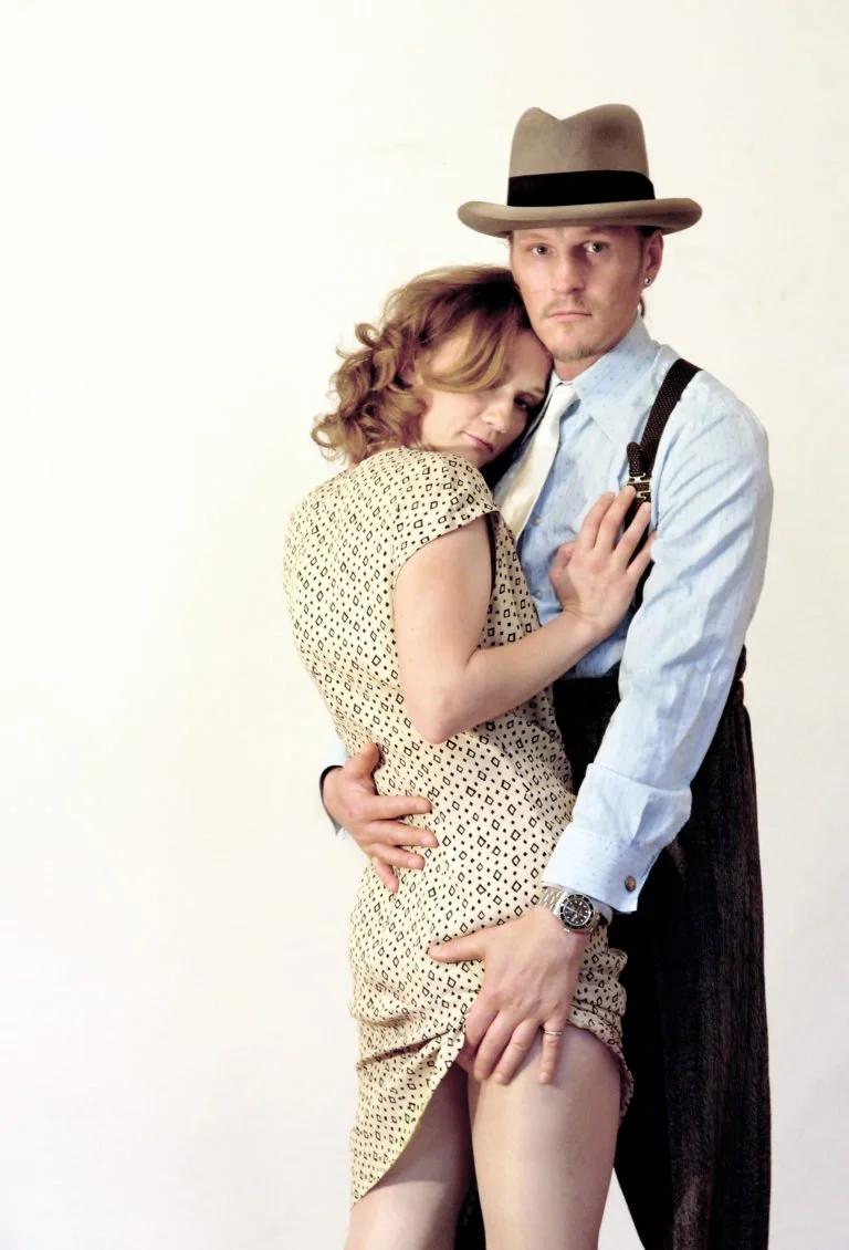 Ein Mann trägt einen braunen Fedora-Hut und ein Hemd, während eine Frau in einem gepunkteten Kleid eng an ihn geschmiegt ist.