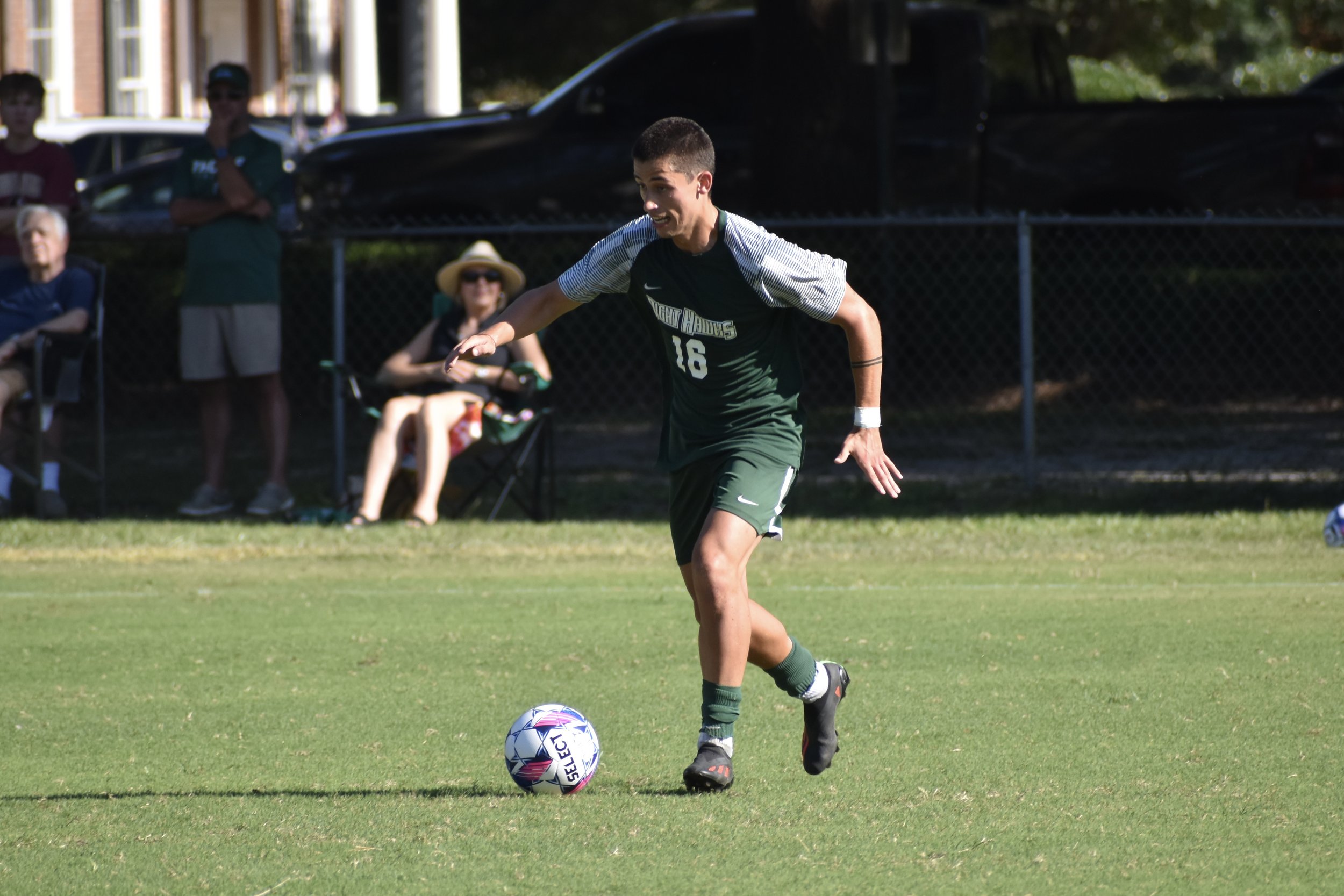 Joven futbolista en uniforme verde y gris, en acción, listo para patear un balón en un campo de césped, con espectadores observando al fondo.