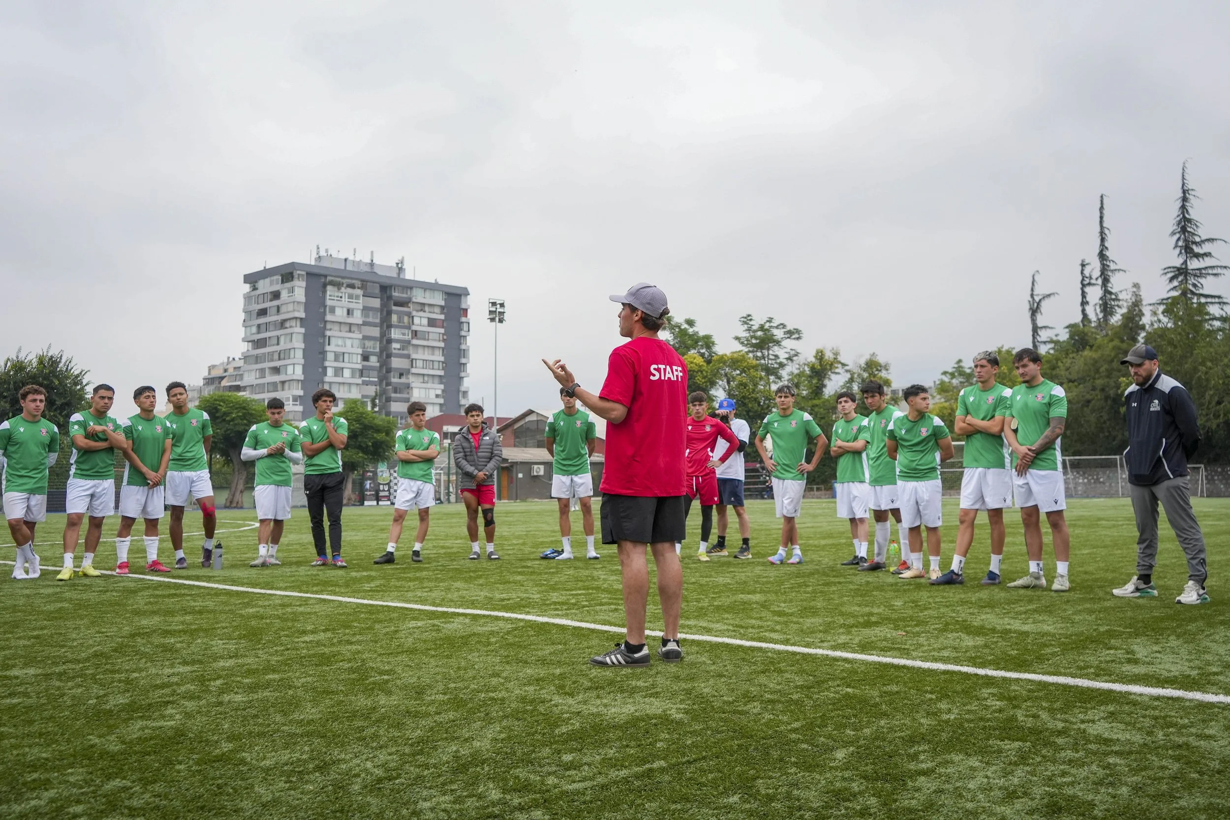 Entrenador hablando a un grupo de jugadores de fútbol en un campo de entrenamiento, todos con uniforme verde y blancos, en un día nublado.